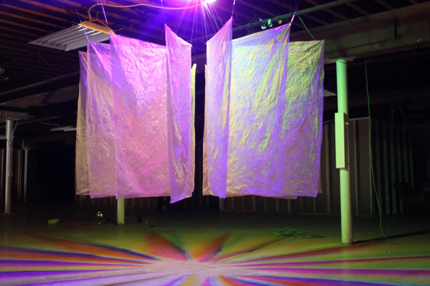 Indoor art installation with large, semi-transparent, iridescent fabric panels hanging from the ceiling, illuminated by colorful lights creating rainbow patterns on the floor.