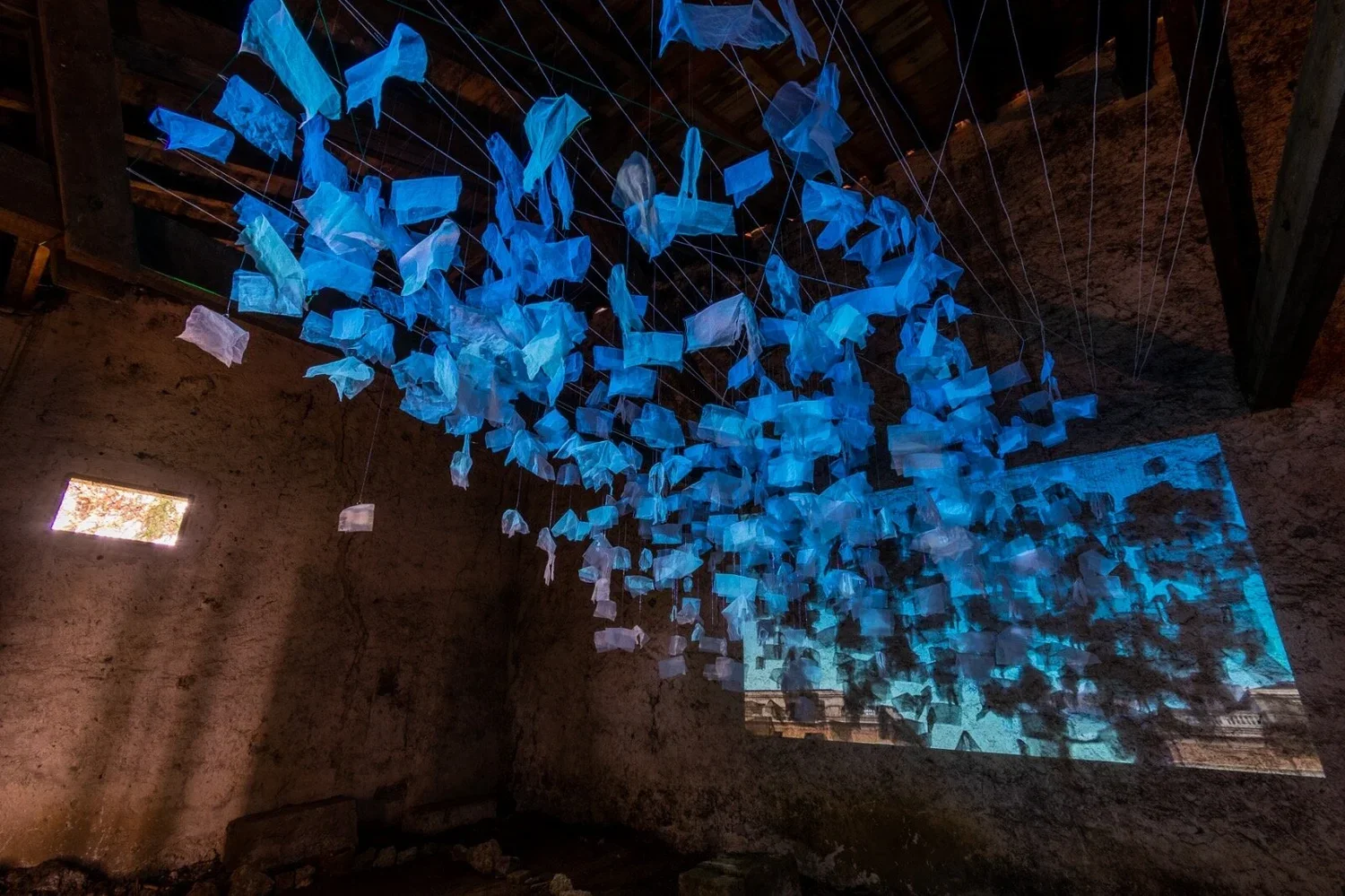 An art installation hanging from the ceiling inside a rustic stone-walled room, consisting of numerous illuminated blue and white paper or fabric pieces suspended with strings.
