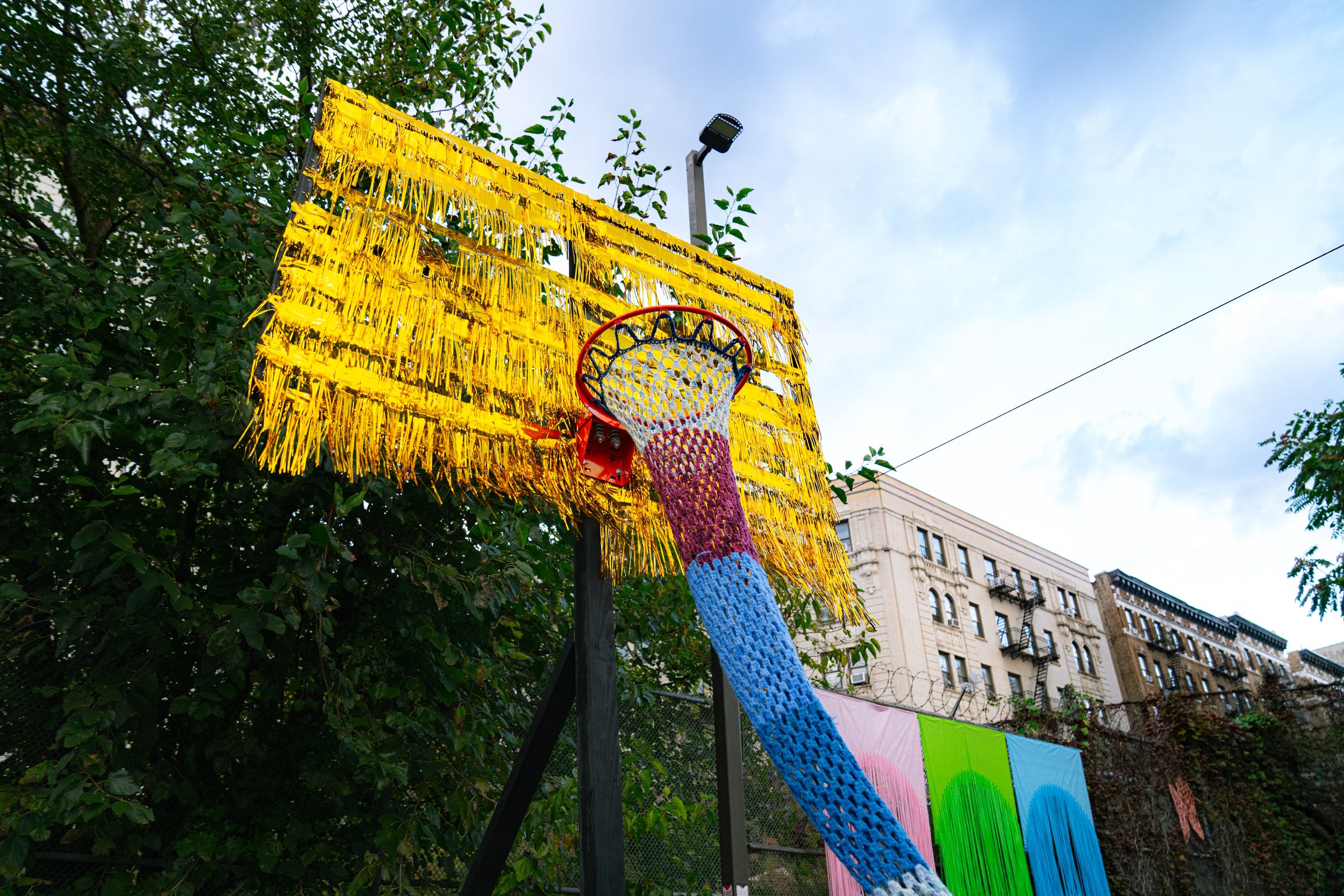Colorful, outdoor basketball hoop with a rainbow in the background, attached to a yellow fringed backboard, and a clear sky.
