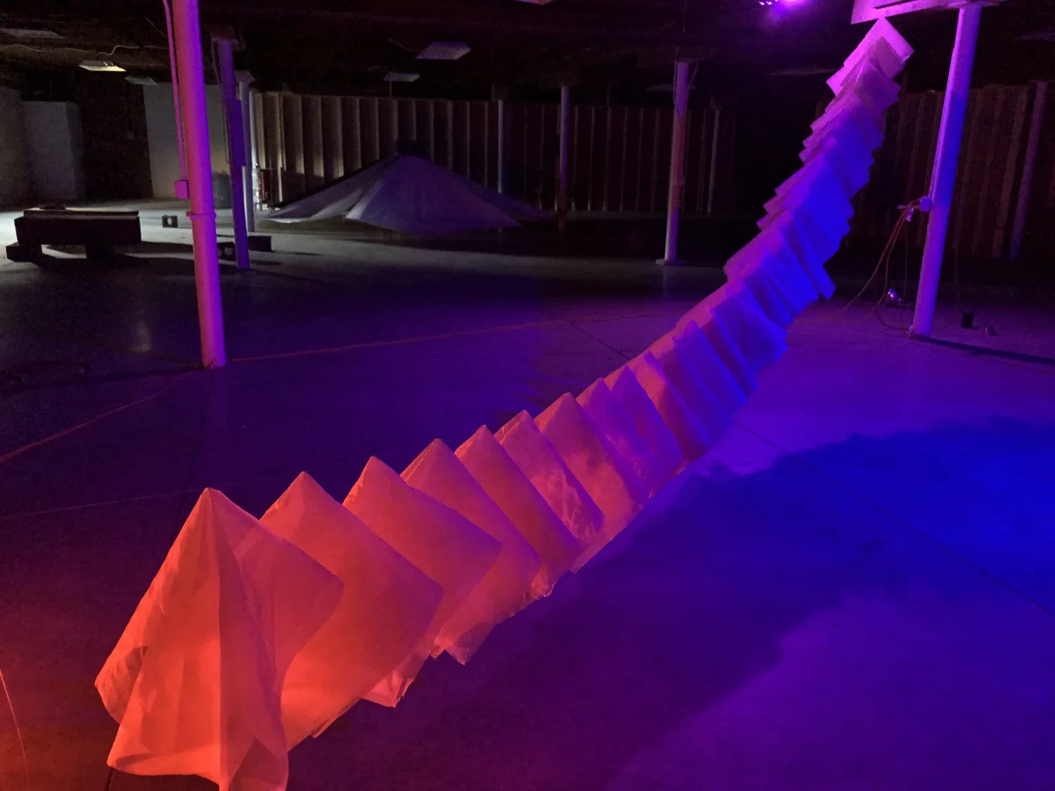 An indoor space with dark lighting illuminated by purple and pink lights, featuring a colorful, abstract sculpture or installation with zigzag shapes.