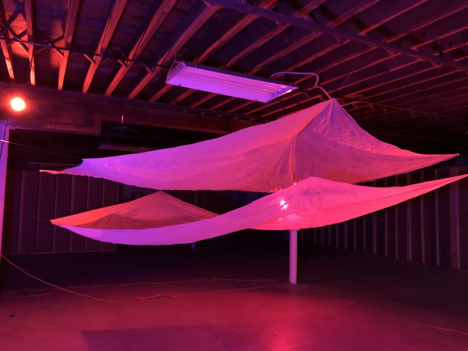 Art installation with three large, pink, fabric-like sails hanging from the ceiling, illuminated by pink and purple lights in a dark room with a wooden ceiling and standing pole.