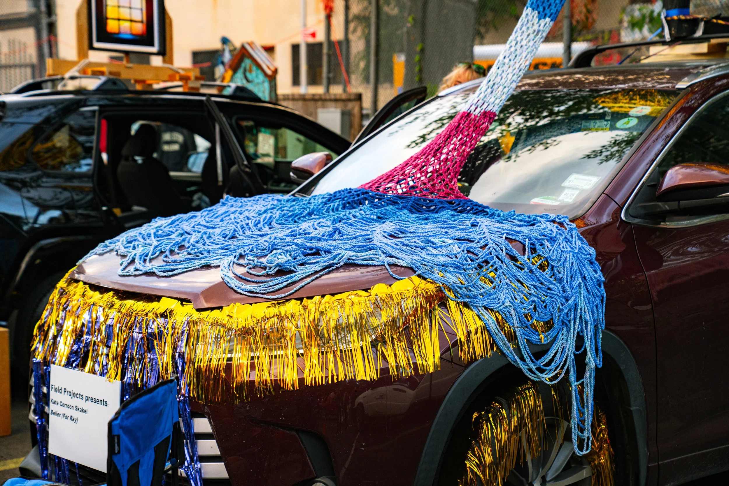 Car decorated with colorful streamers and yarns, likely for a parade or celebration, with a sign indicating it is part of a presentation by Field Projects.