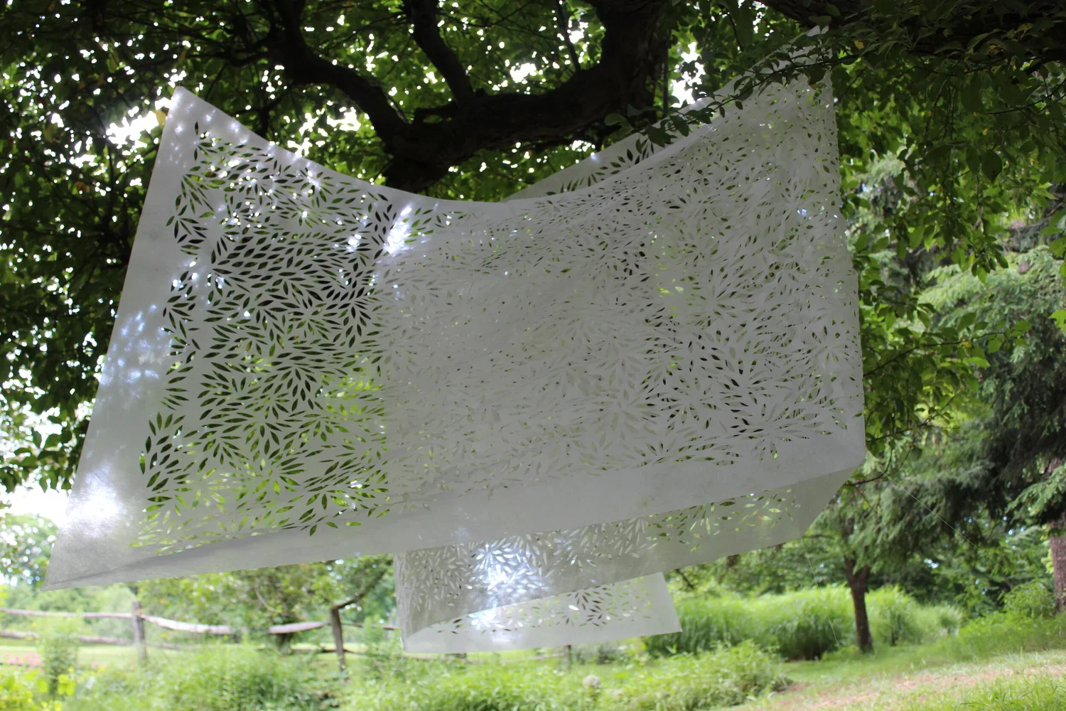 White decorative paper sheets with intricate floral cutout patterns hanging from a tree branch in a green outdoor park.