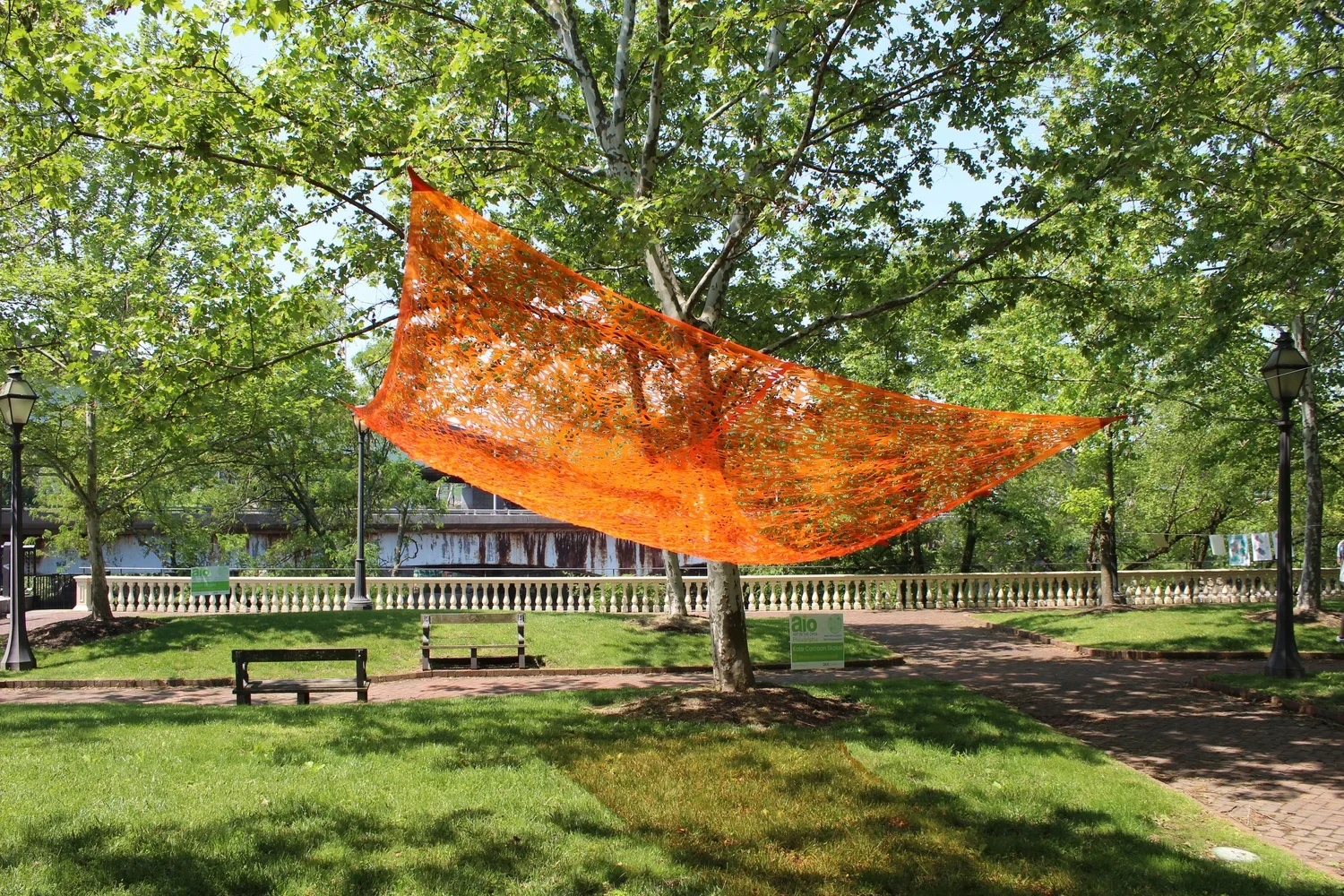 A large orange netted hammock hanging between two trees in a park with green grass, benches, and vintage-style street lamps.