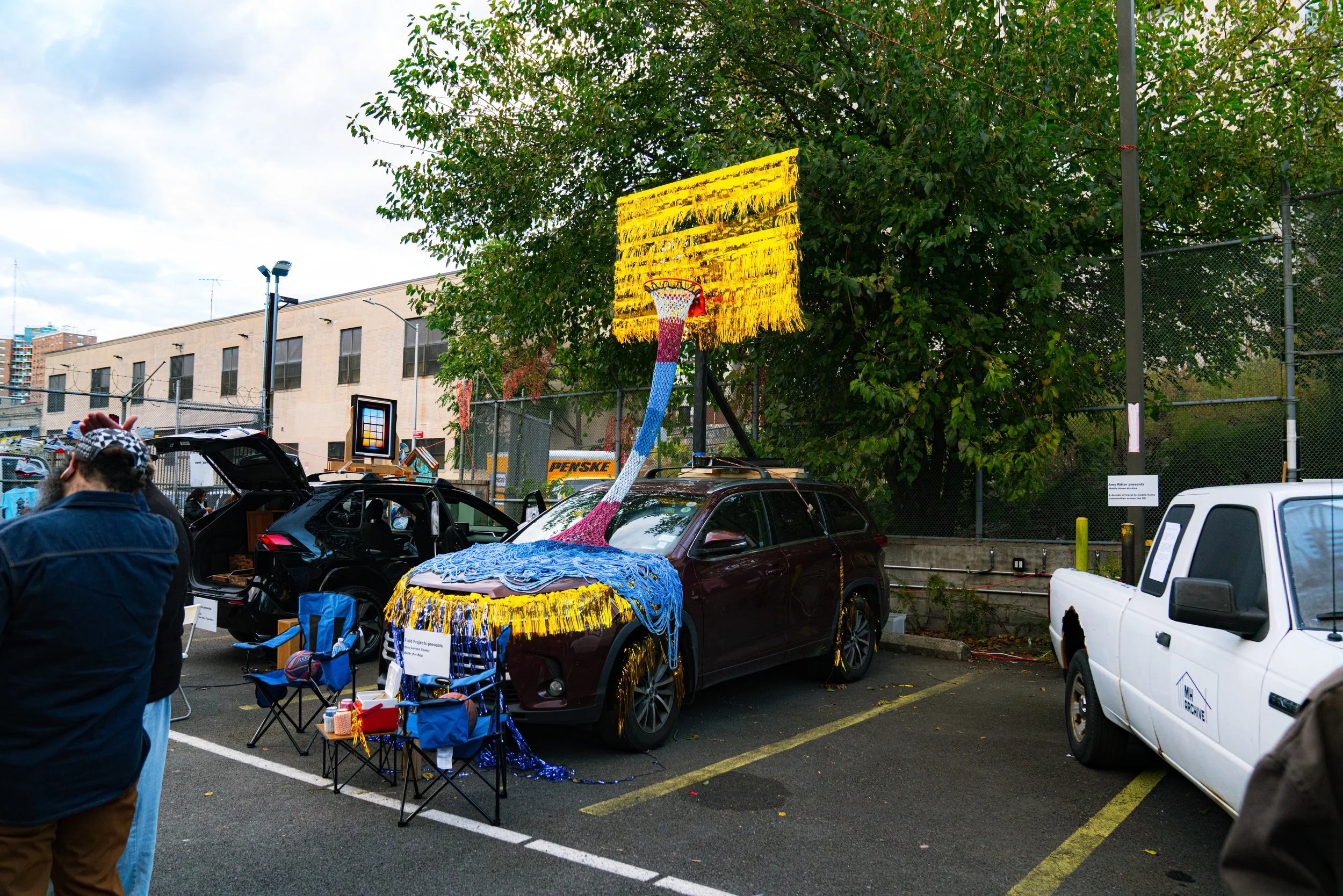 A parking lot with a dark red car decorated as a basketball with a hoop on top and a backboard, presentaing a creative art installation.