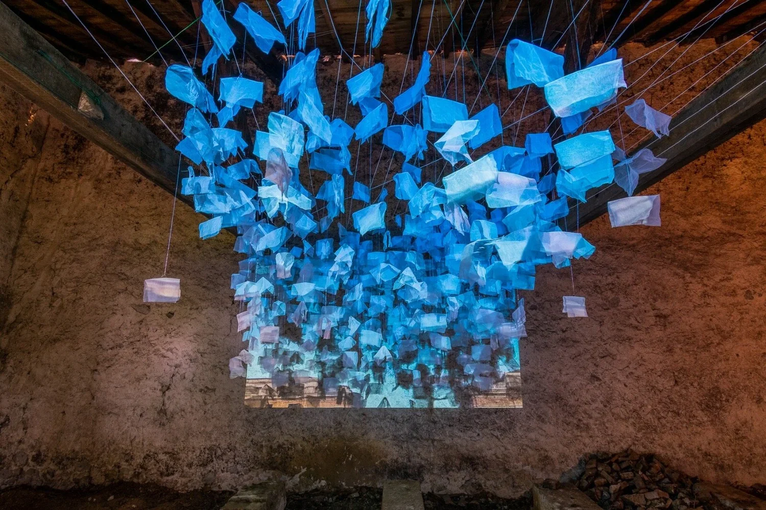 An art installation with numerous hanging blue and white fabric pieces suspended from the ceiling inside a rustic, stone-walled room.