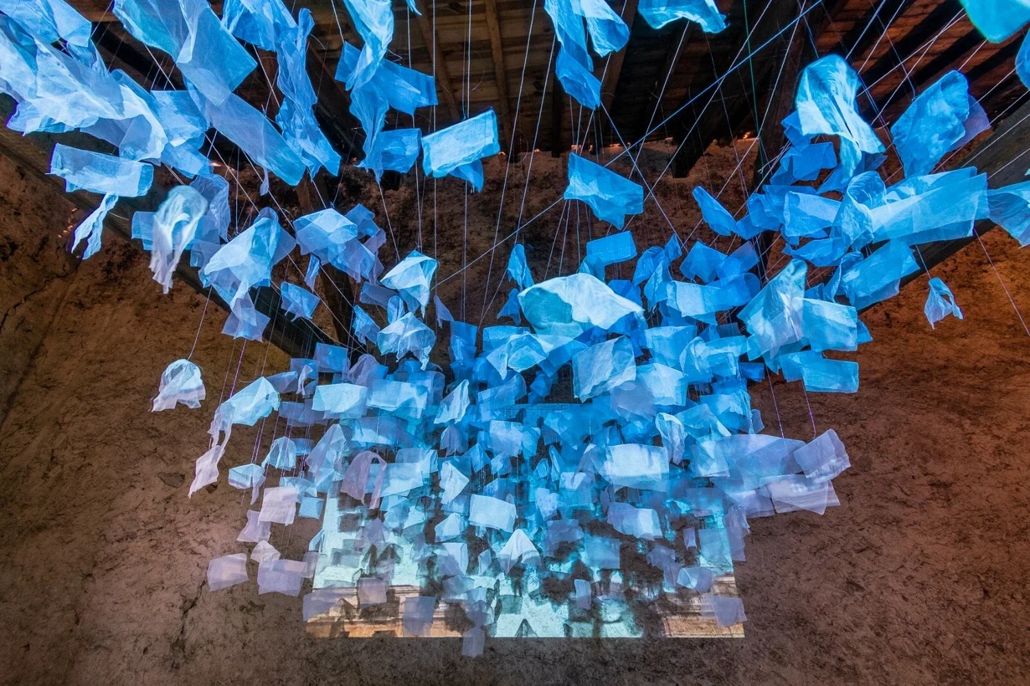An art installation with hanging blue and white fabric pieces suspended from thin strings, creating a cloud-like appearance inside a rustic space with stone walls and a wooden ceiling.