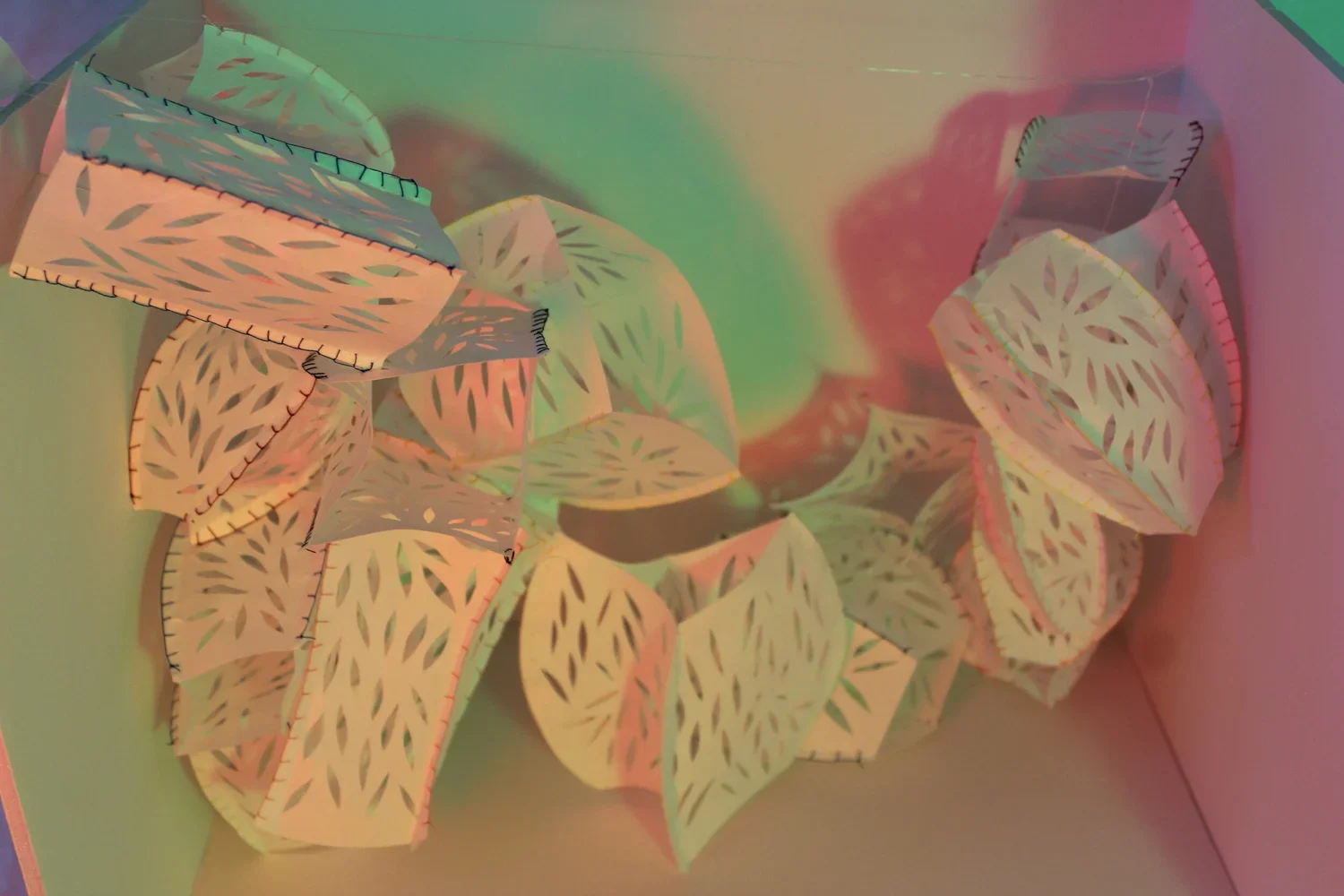 Paper lanterns with leaf cutout designs hanging inside a box, illuminated with colorful pastel lighting.