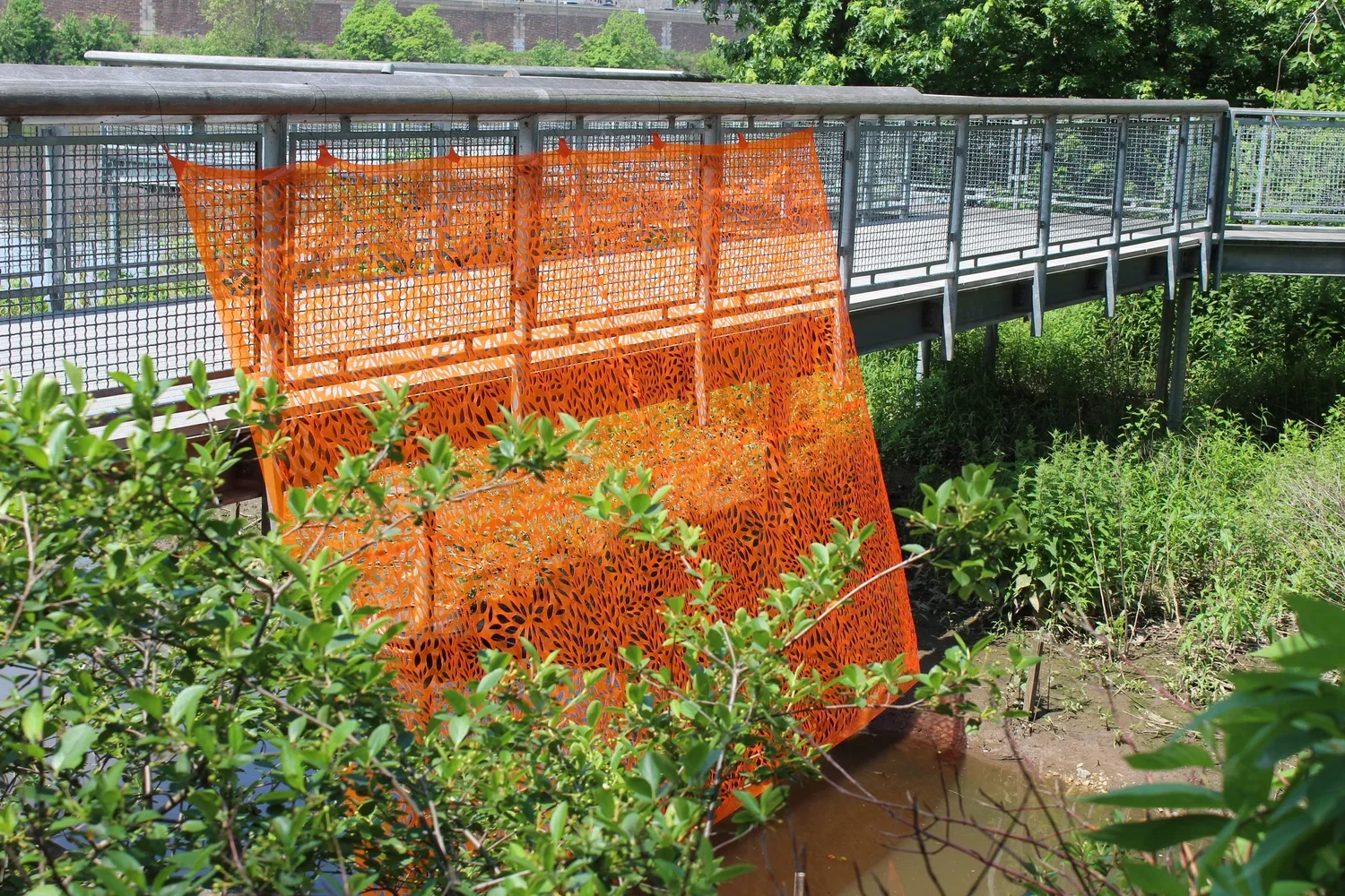 A bridge with a railing over a green, leafy area with a small waterway below. Orange safety barriers with a cut-out leaf pattern are attached to the railing, possibly for construction or maintenance.