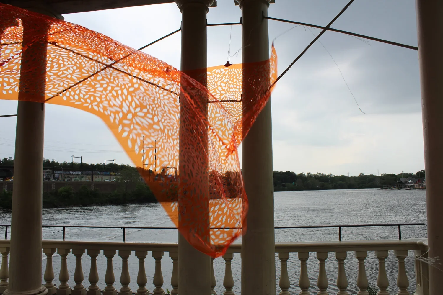 Orange patterned fabric draped over white pillars overlooking a river with a railing, overcast sky, and distant trees.