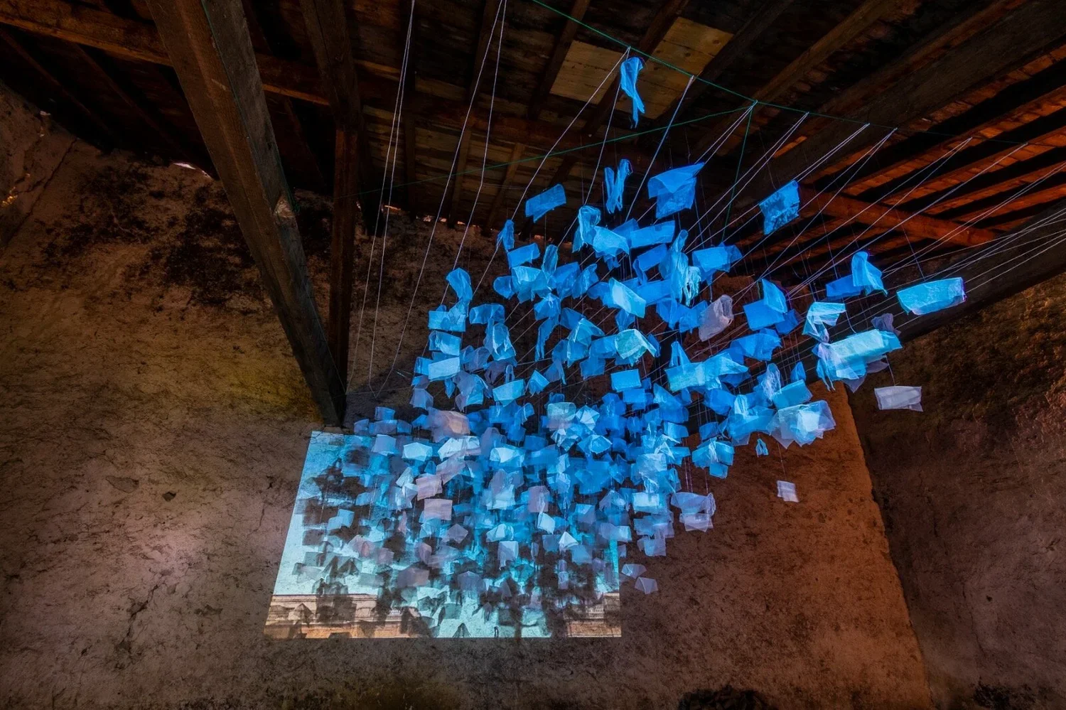 Installation art featuring numerous blue fabric pieces hanging from strings in a rustic, dimly lit space with textured walls and wooden ceiling beams.