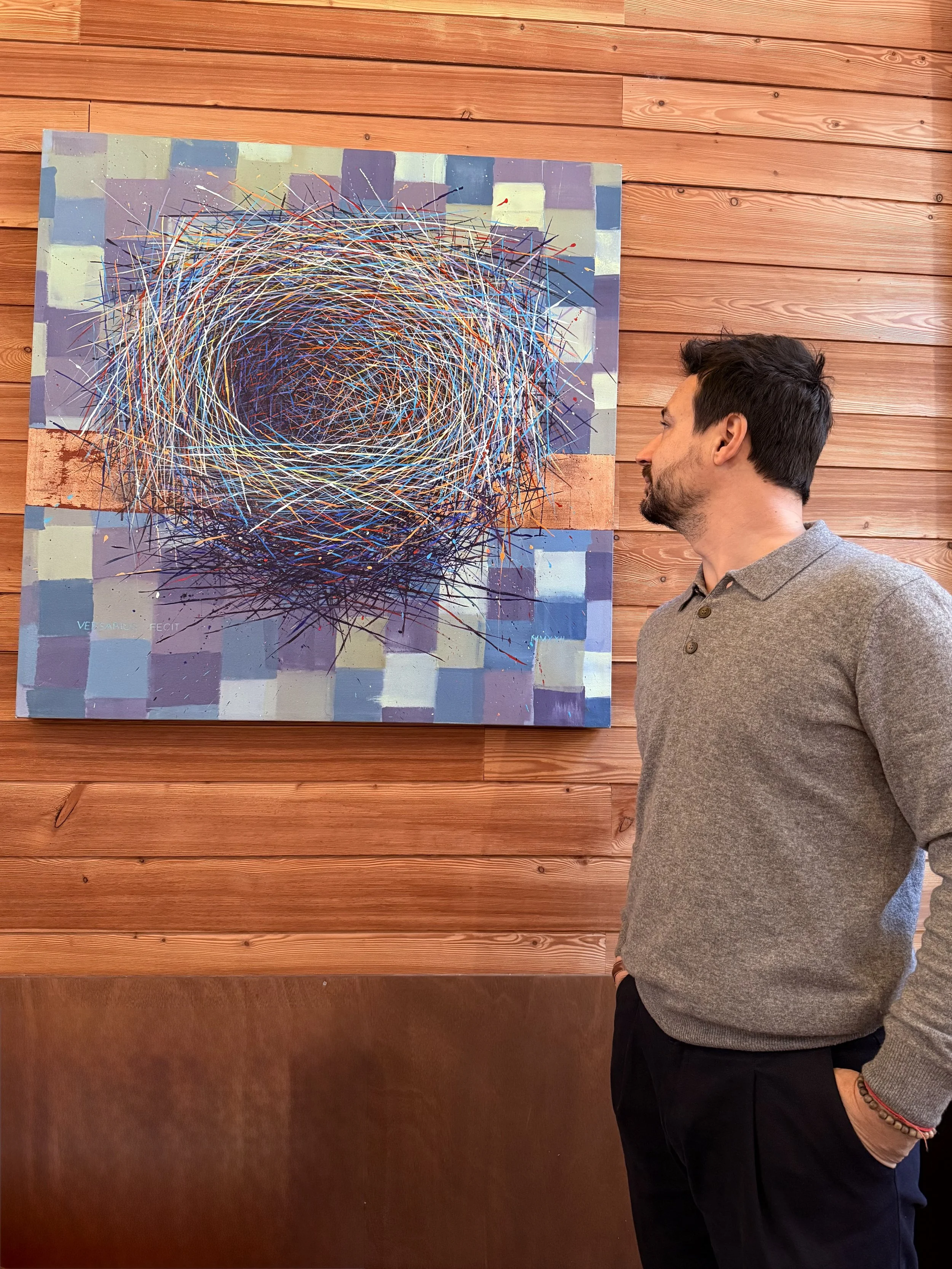 Uomo in piedi che osserva un quadro astratto con linee colorate e sfondo geometrico, appeso a una parete di legno.