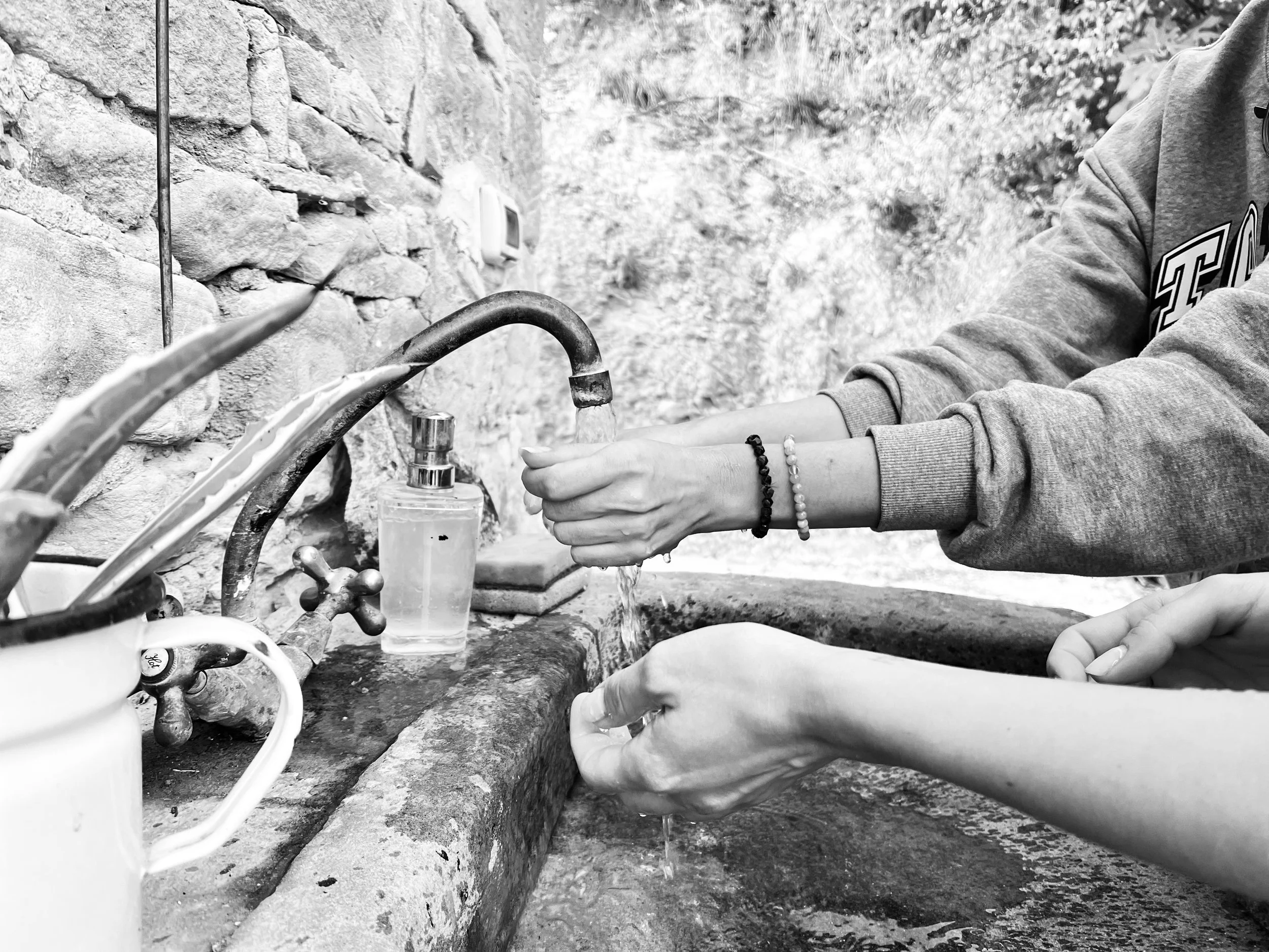 Due persone lavano le mani sotto un rubinetto esterno, con una bottiglia di sapone liquido accanto, in una zona con pareti in pietra.