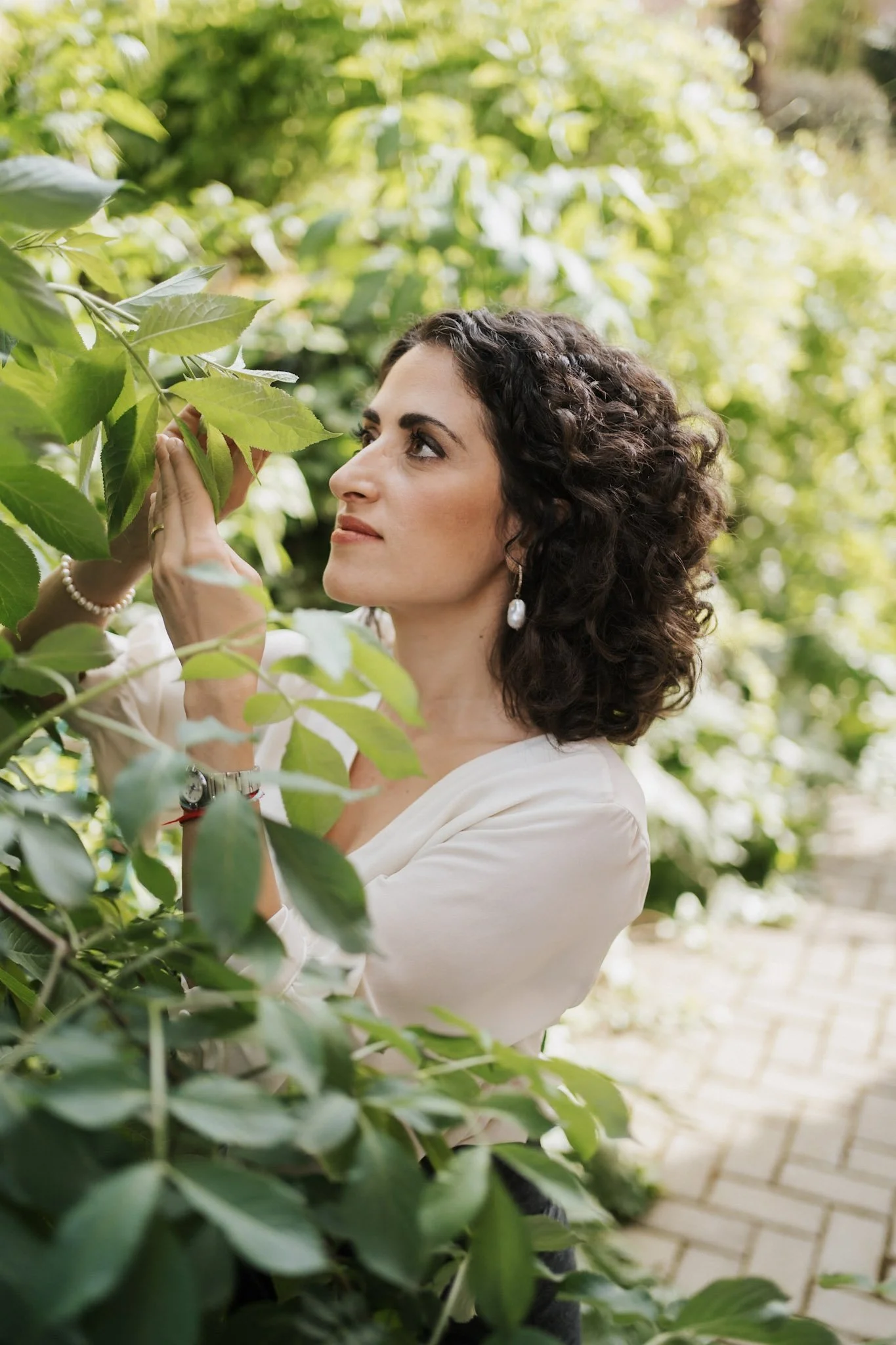 Una donna con capelli ricci scuri e orecchini di perle si trova tra foglie verdi, che osserva attentamente, in un ambiente naturale soleggiato.