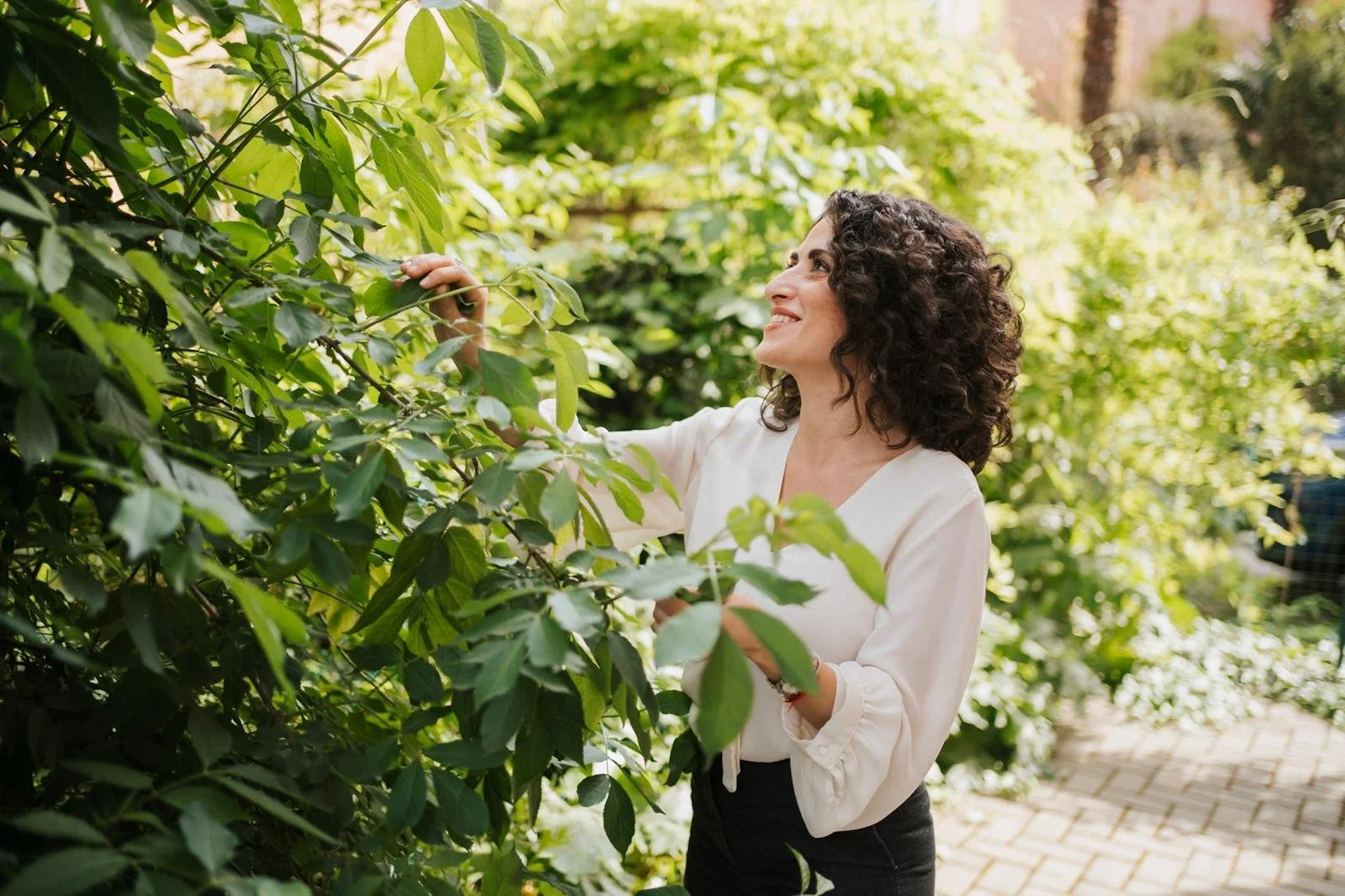 Una donna con capelli ricci scuri indossa una maglietta bianca e si trova in un giardino circondata da vegetazione verde, sorride mentre tocca le foglie di una pianta.