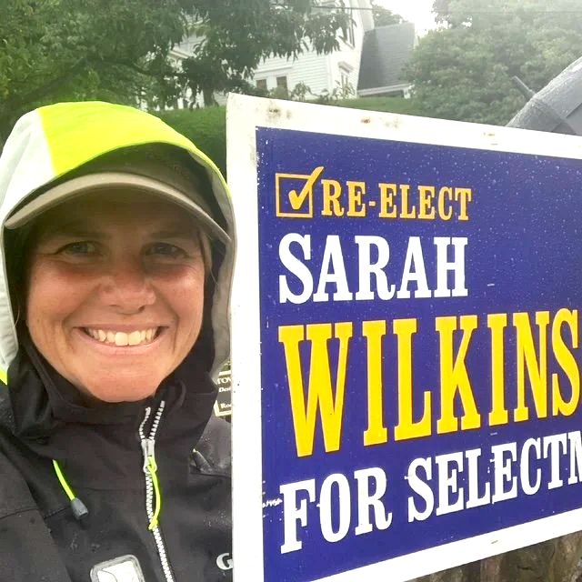 A woman smiling outdoors, wearing a yellow and gray hooded rain jacket, standing next to a political campaign sign that reads 'Re-elect Sarah Wilkinson for Selectman'.