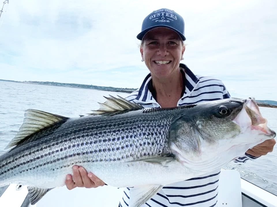 Woman in a striped shirt and a blue cap holding a large striped bass fish on a boat on the water, smiling.
