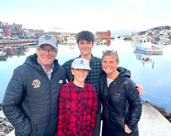 A family of four smiling and posing by the harbor, with boats and buildings in the background.