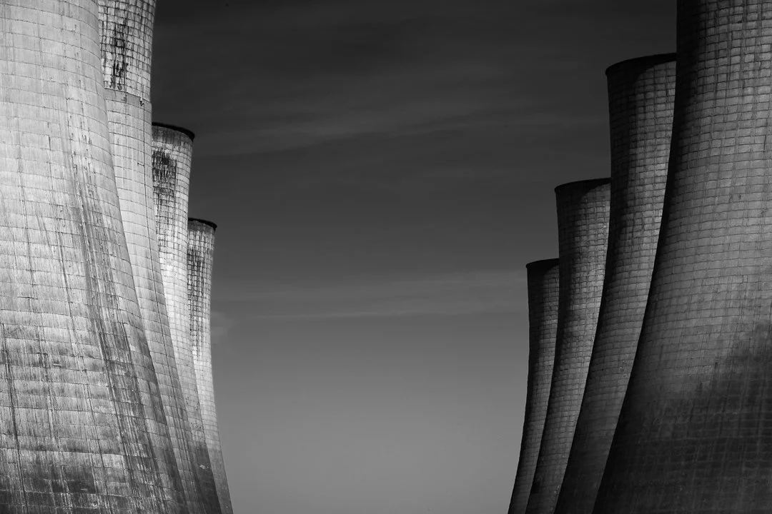 Monumental concrete cooling towers rise against a dark sky, their curved surfaces and weathered textures highlighted by sunlight.