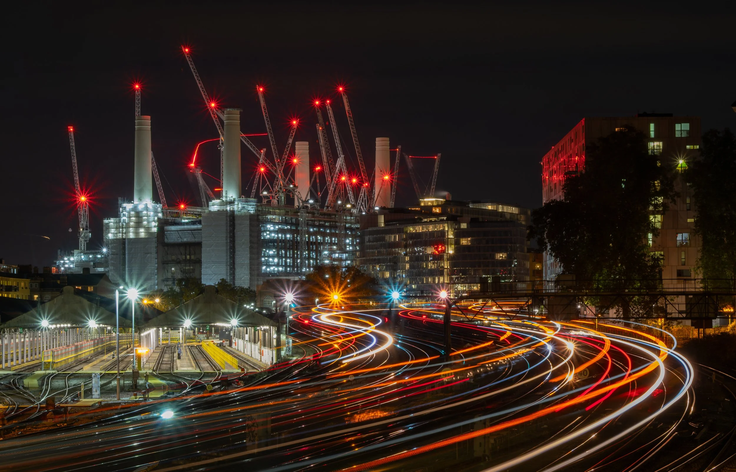 Battersea Power Station seen at night from Ebury Bridge, surrounded by construction cranes and sweeping train light trails.