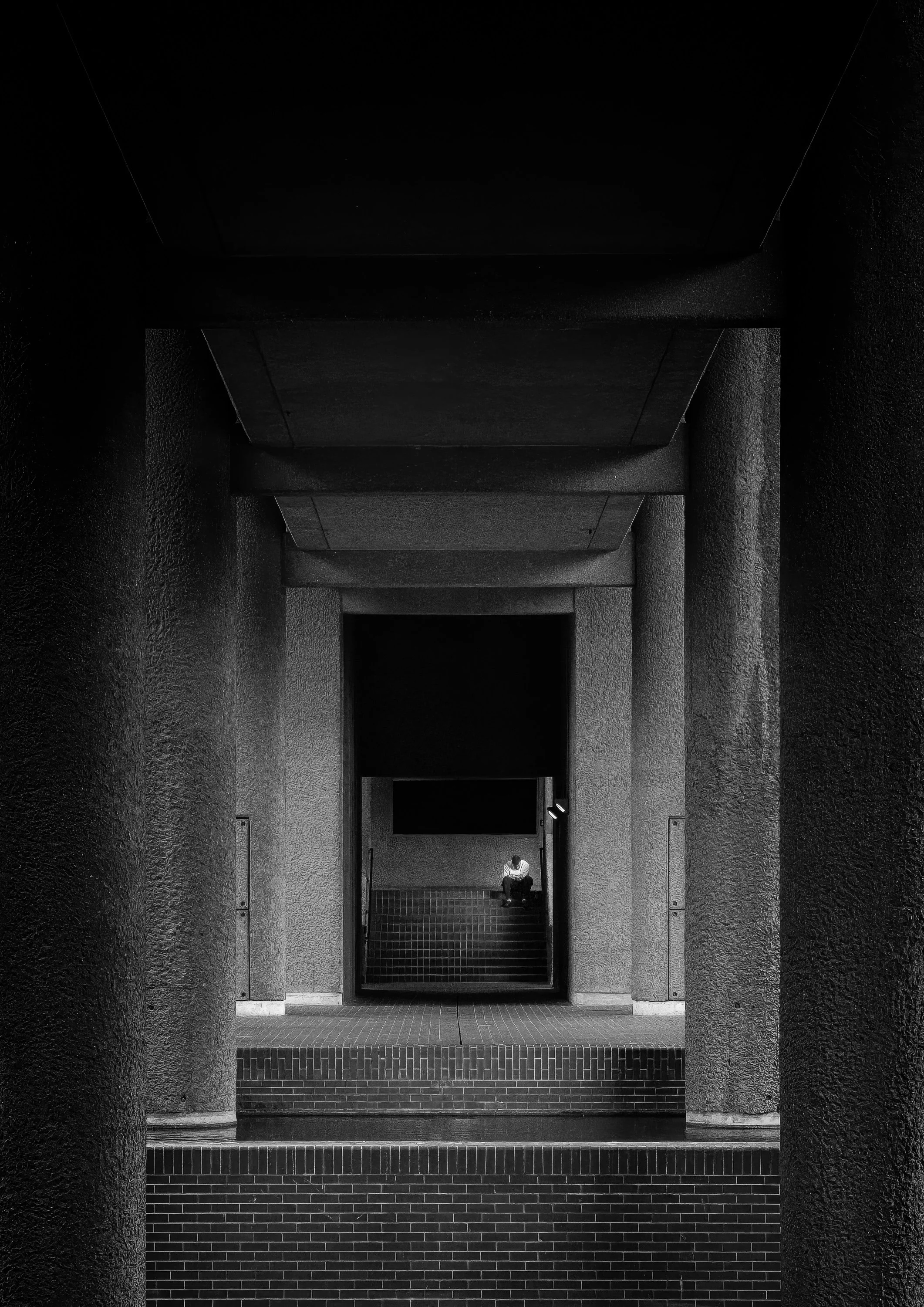 Lone figure seated at the end of a concrete corridor in the Barbican Estate, framed by repeating rectangular openings.