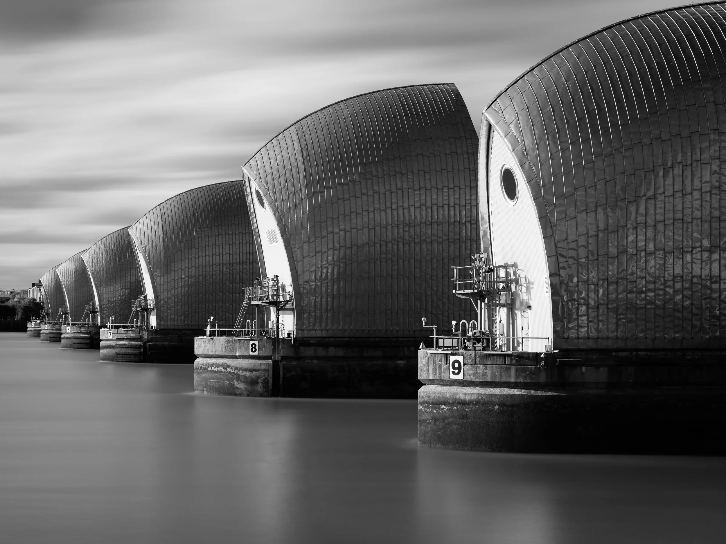 Thames Barrier piers stretching across the river beneath a cloudy sky, reflected in smooth water.