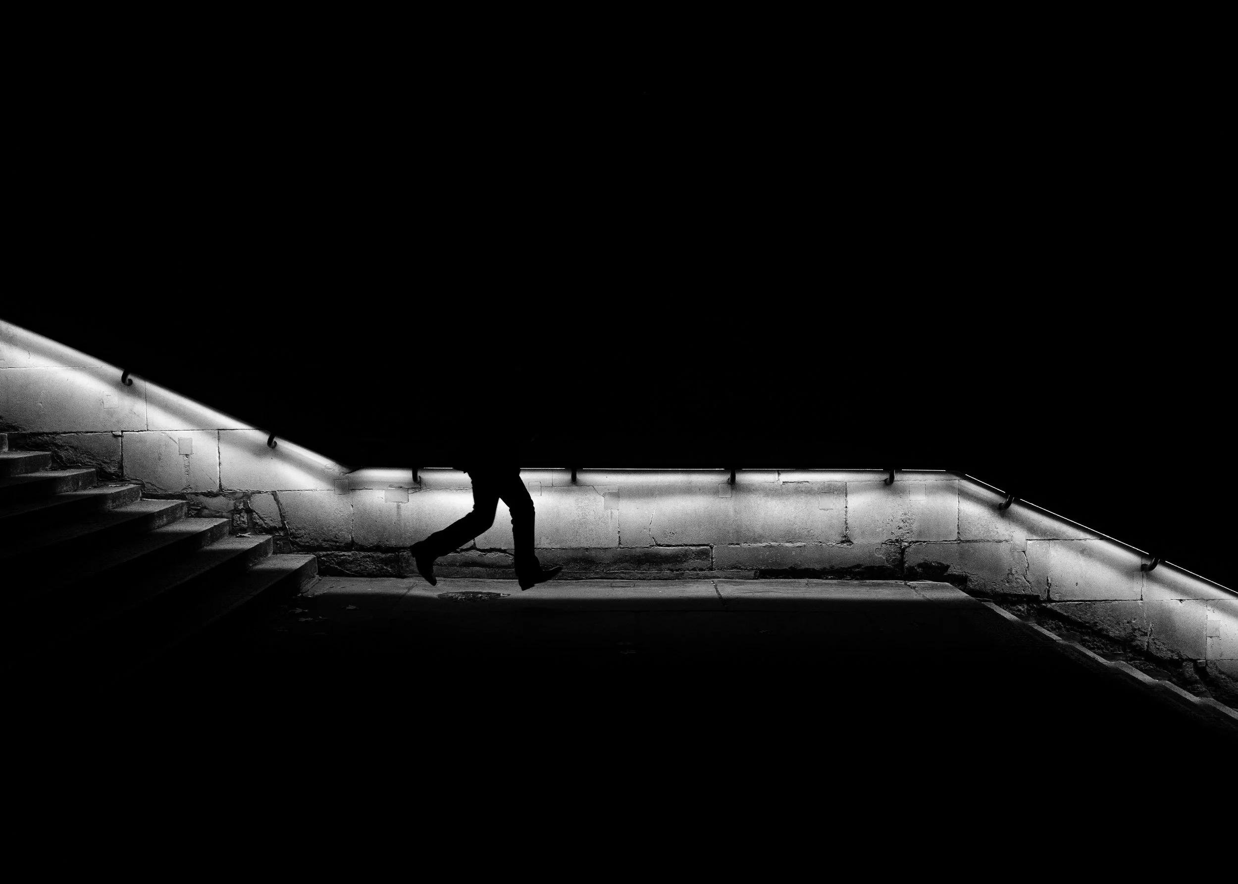 Lone figure walking along a curved illuminated stairway in St James’s, surrounded by deep shadow.