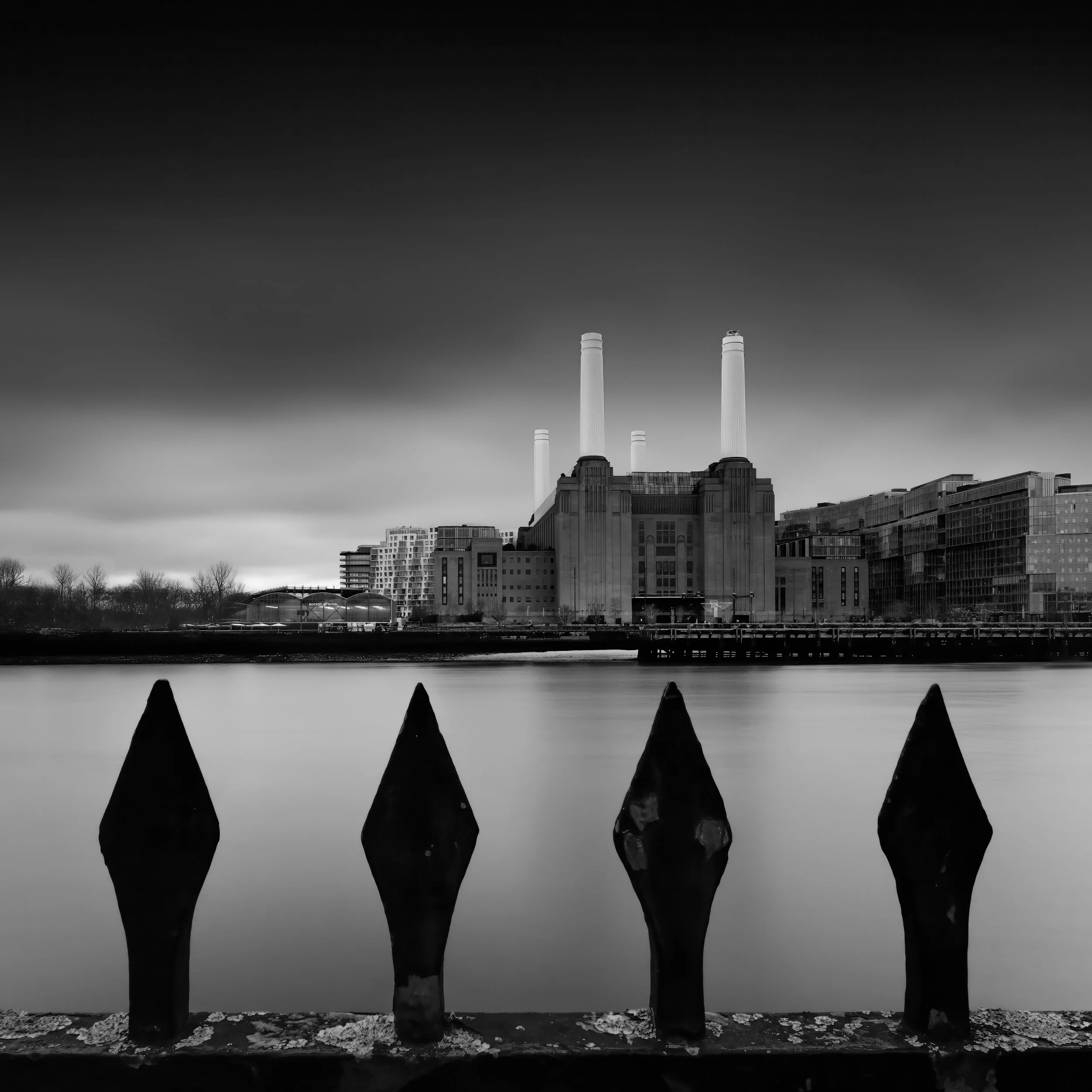 Battersea Power Station viewed across the Thames from Chelsea Embankment, framed by pointed riverside railings in the foreground.