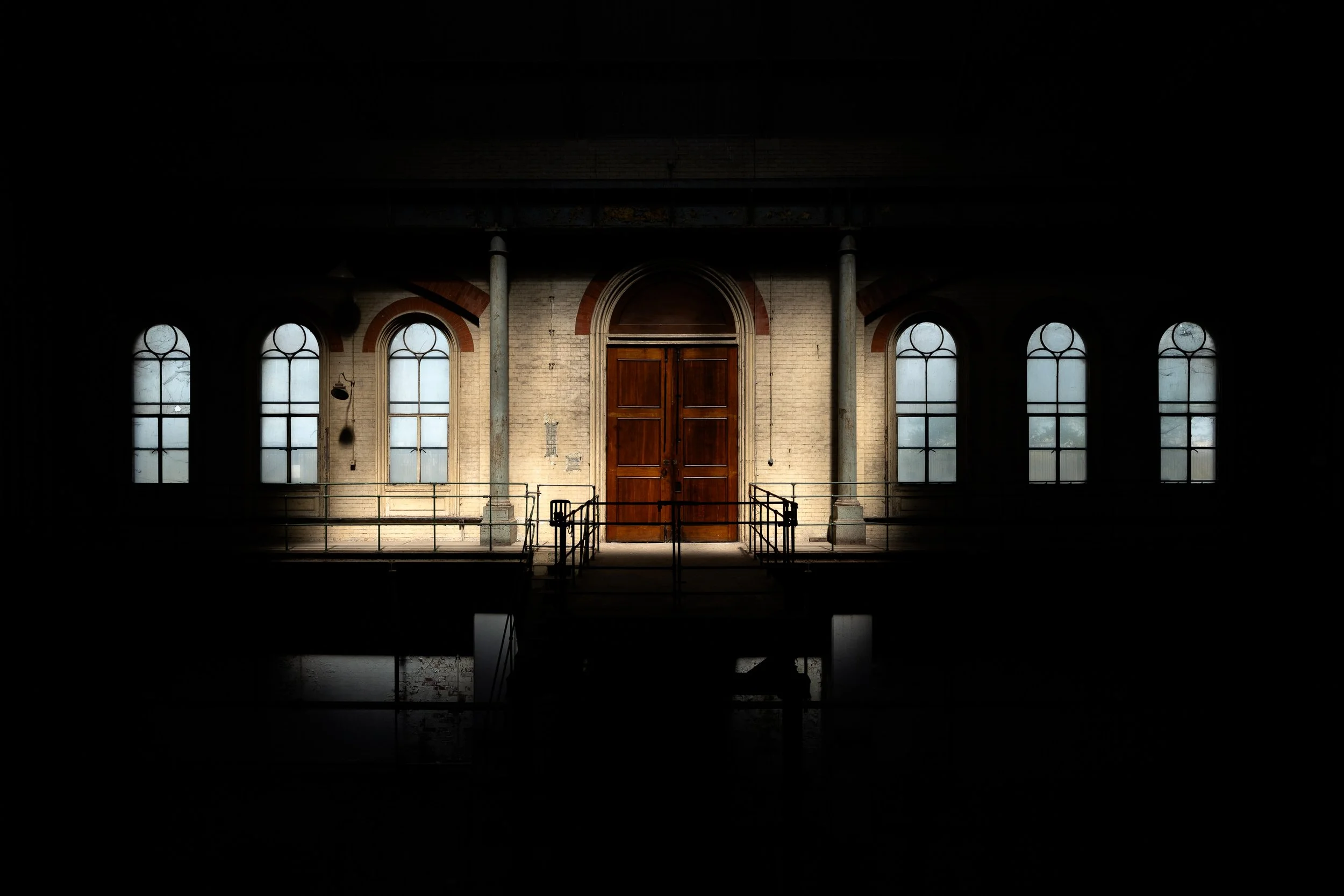 Dimly lit interior of Crossness Pumping Station, with tall windows, brickwork and a central doorway picked out by natural light.