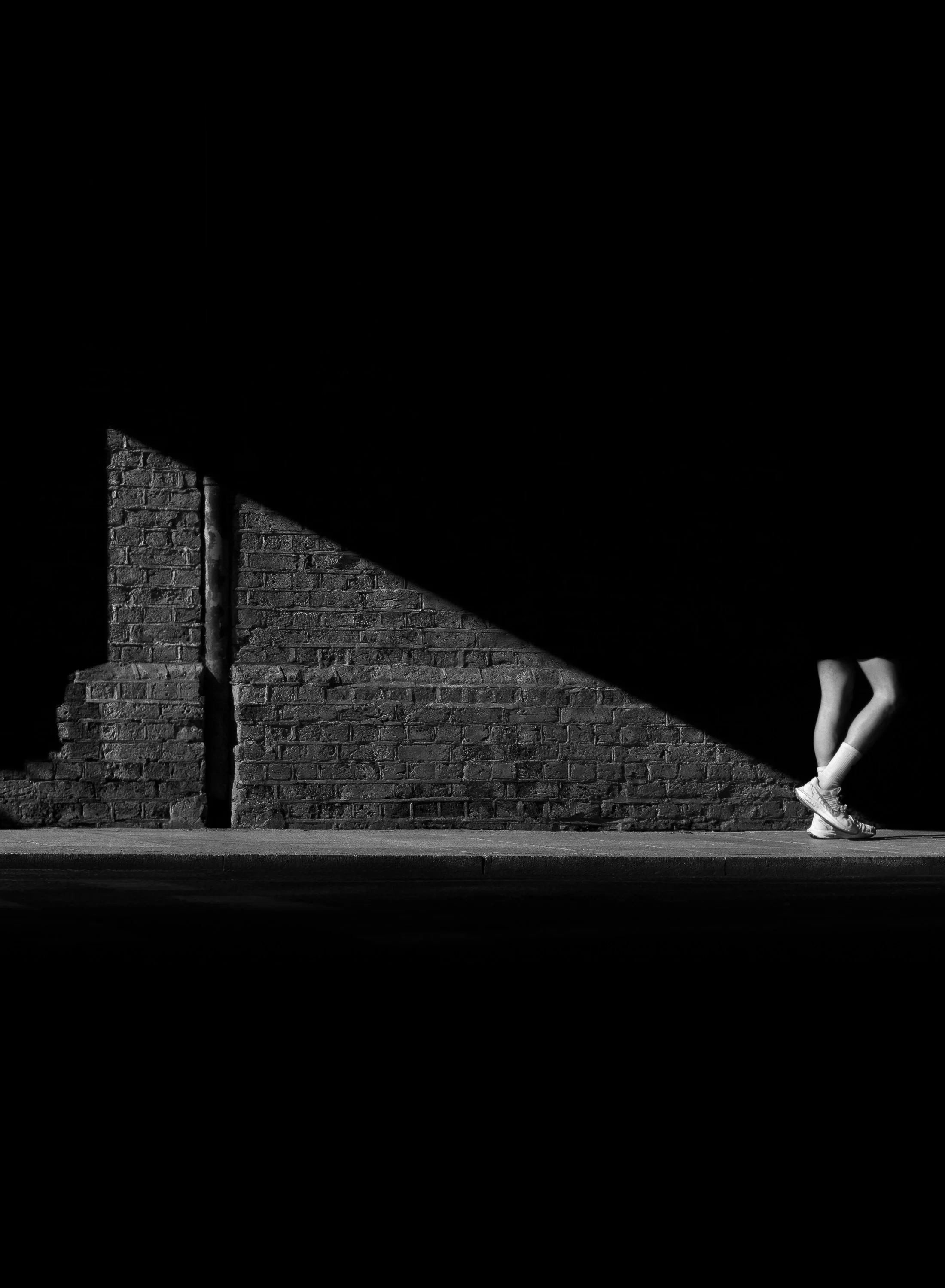 Person in white trainers standing below a railway bridgel in Wandsworth Town, crossed by a strong triangular shadow.