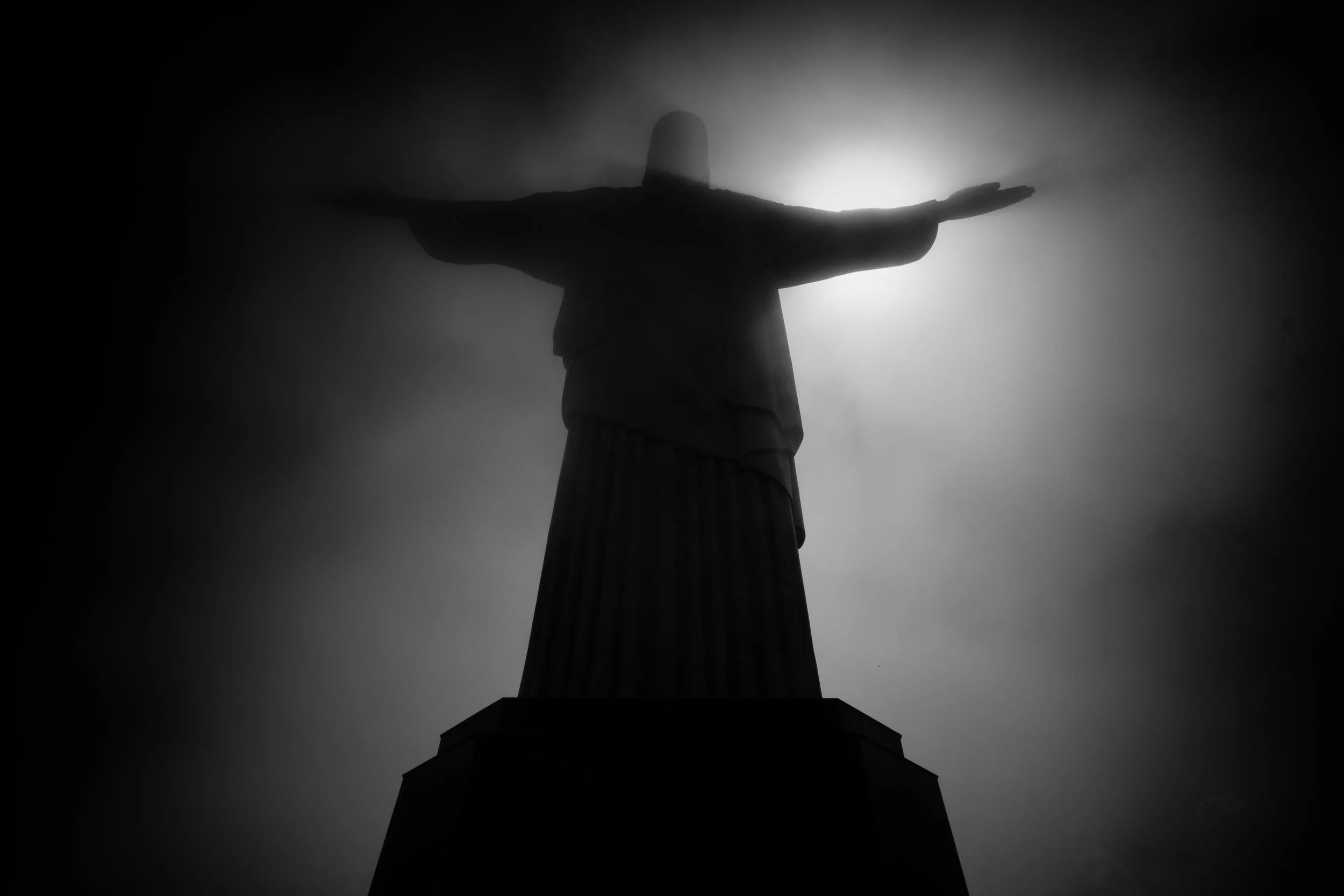 Christ the Redeemer silhouetted against a bright, clouded sky in Rio de Janeiro, viewed from below.