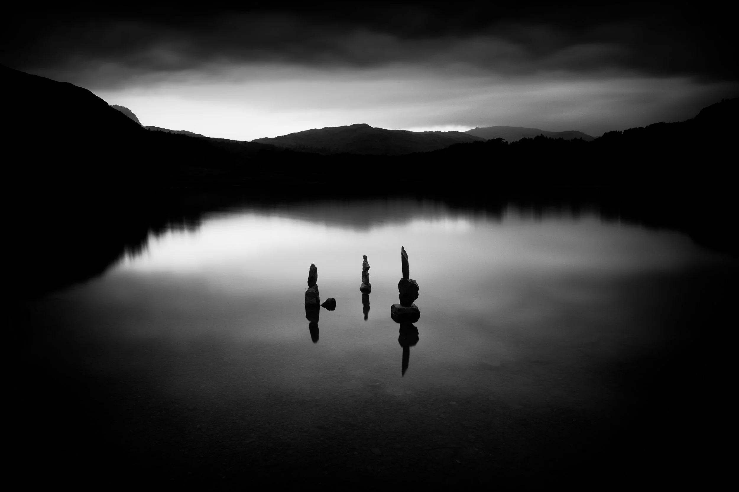 Rydal Water in the Lake District, with stacked stones in the foreground and distant hills reflected in the calm water.