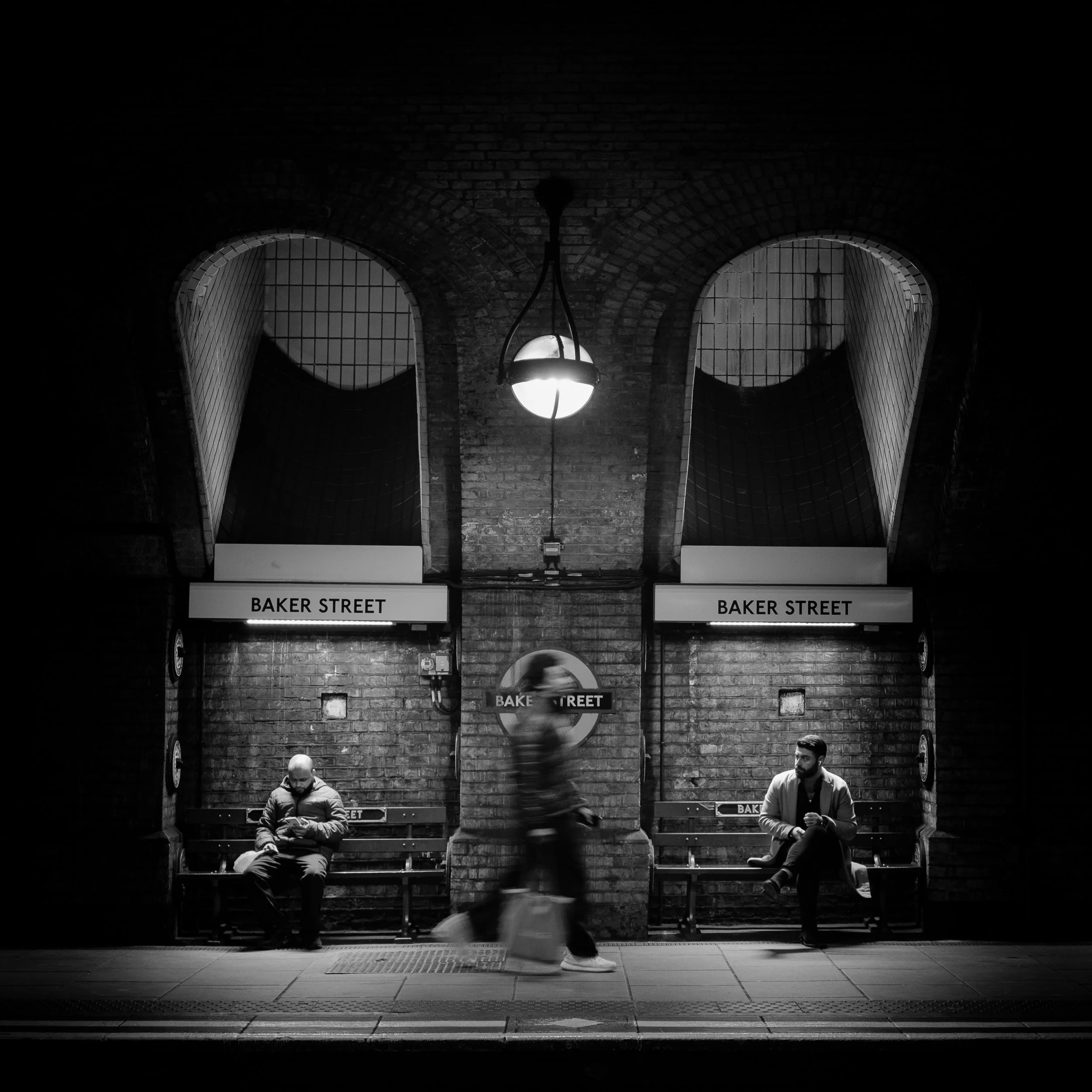 Baker Street Underground station platform, with a blurred passer-by moving beneath the station signs and hanging lights.