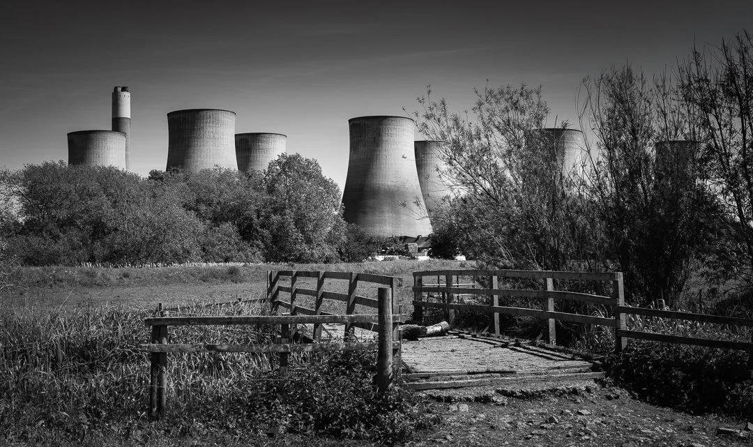 A rural footpath and wooden fence lead towards trees partially concealing the cooling towers of Ratcliffe-on-Soar Power Station.