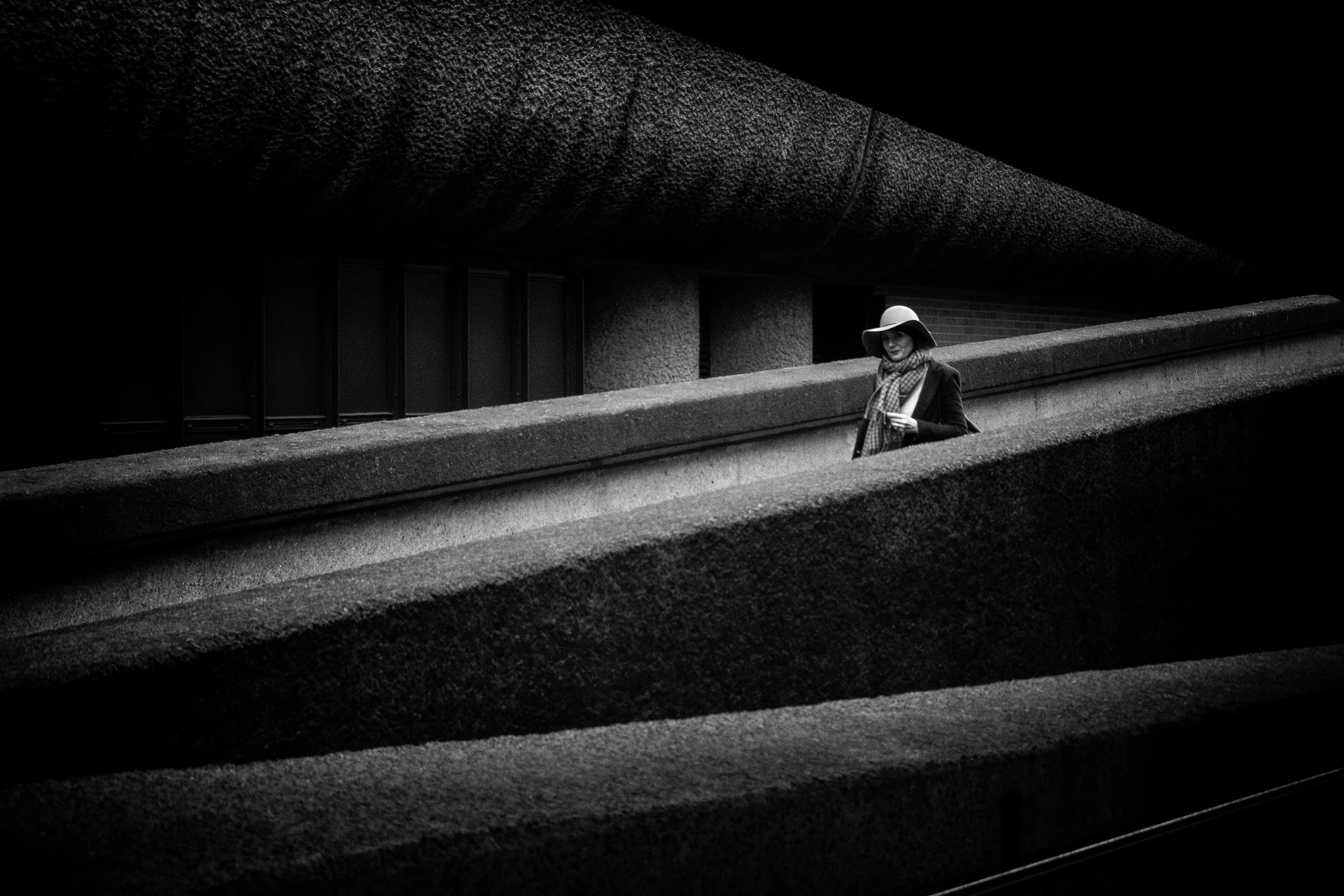 Woman standing on the stepped concrete terraces of the Barbican Estate, framed by strong horizontal lines and deep shadow.
