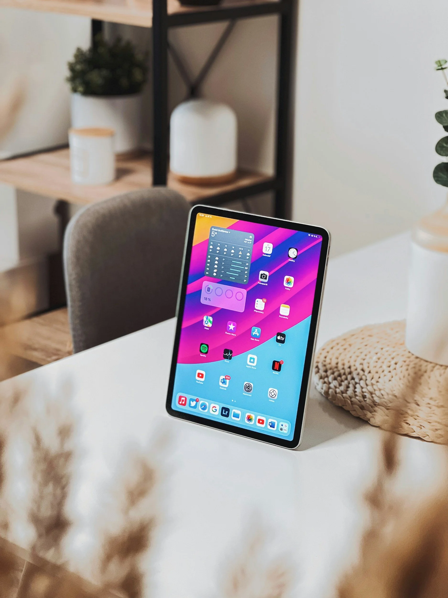 An iPad with a colorful home screen standing on a white table in a cozy room with a beige woven placemat, a white vase, and green plants.
