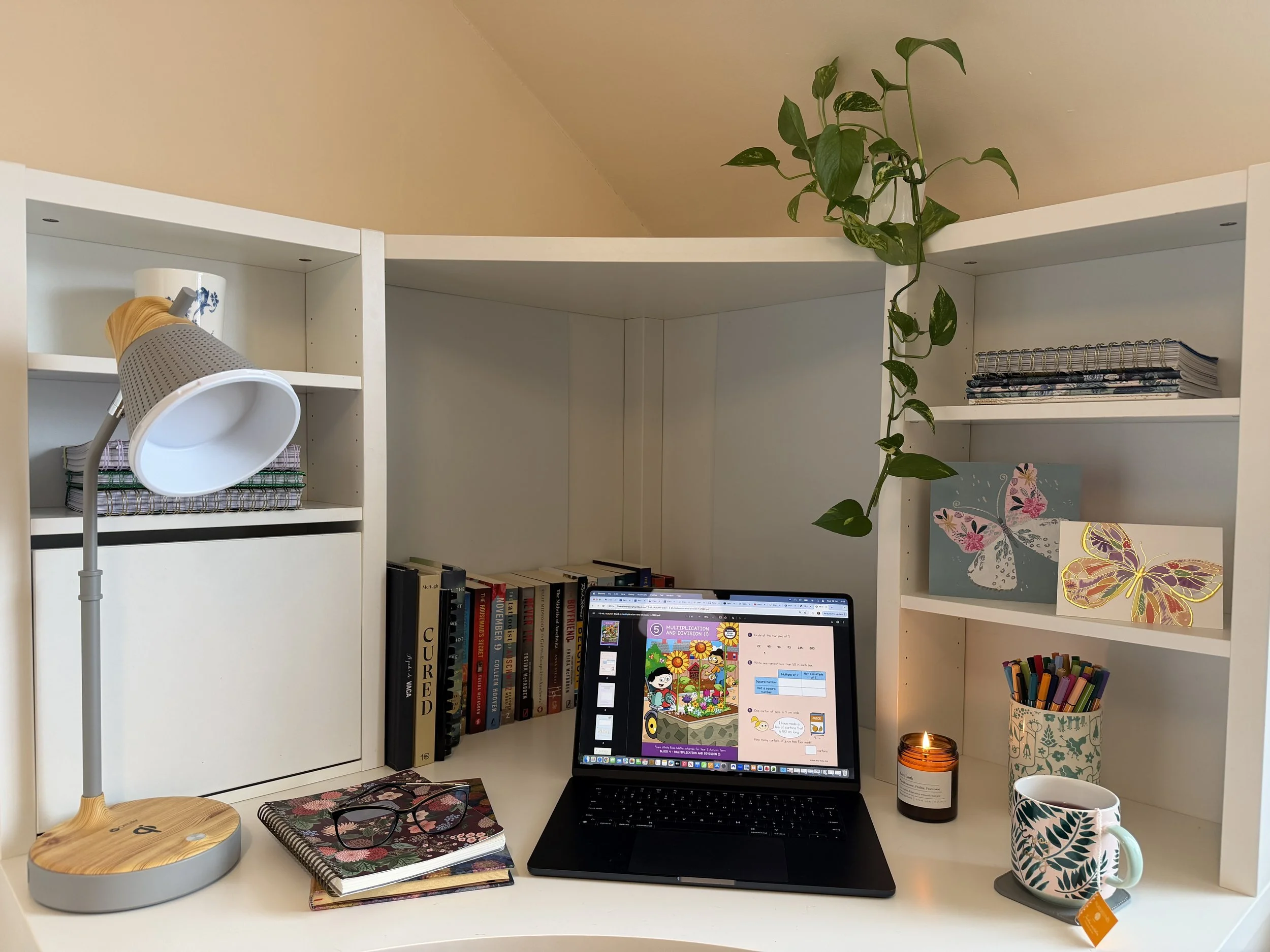 A neatly organized white desk workspace with a laptop, books, art, and a lamp, decorated with a hanging plant and artwork, and a lit candle and mug.