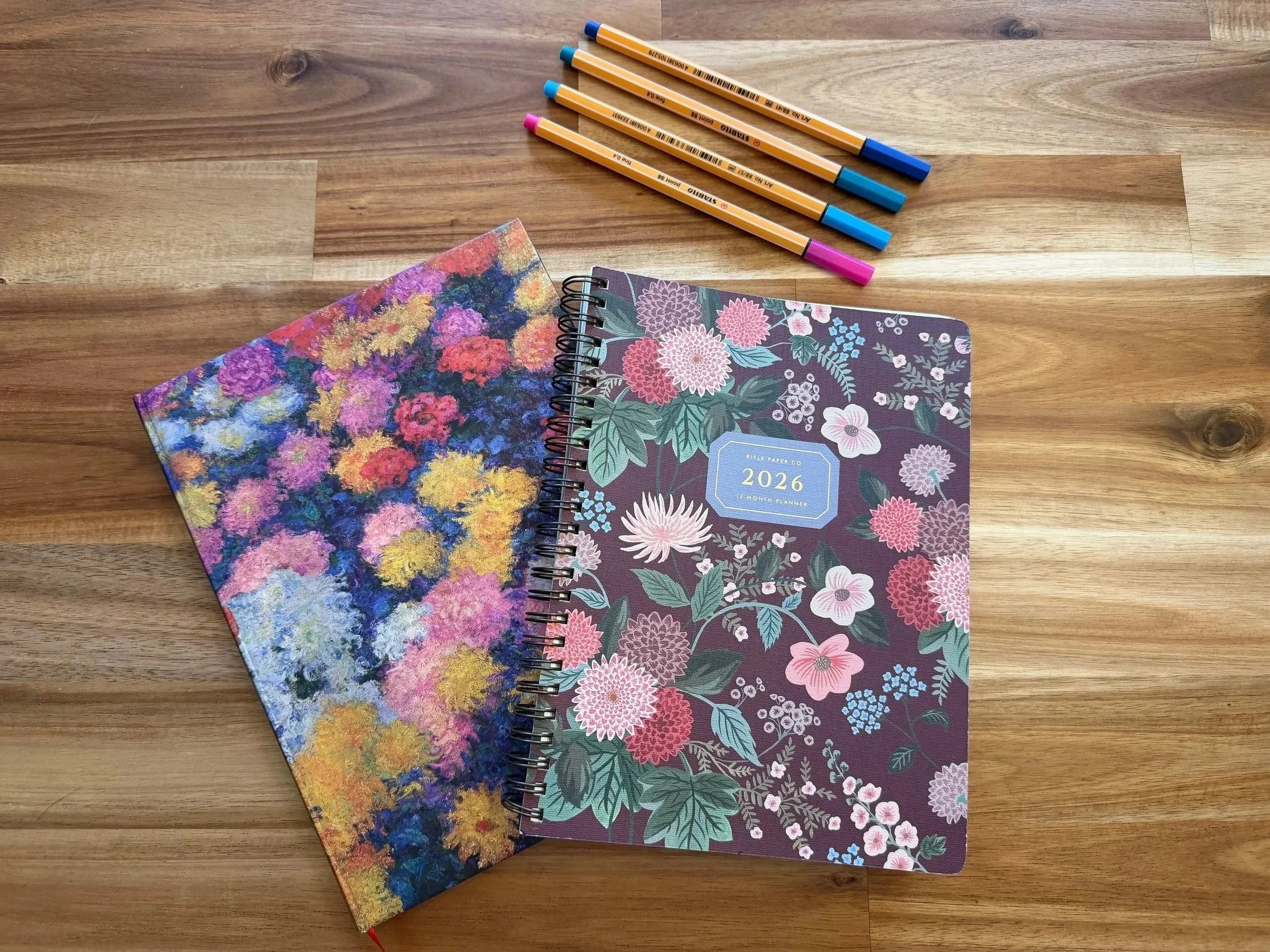 A wooden desk holds two floral patterned 2026 planners, one with a dark purple cover and pink, white, and green flowers, and the other with an impressionist-style multicolored flower painting. Four colored pens with blue and pink caps are above the planners.