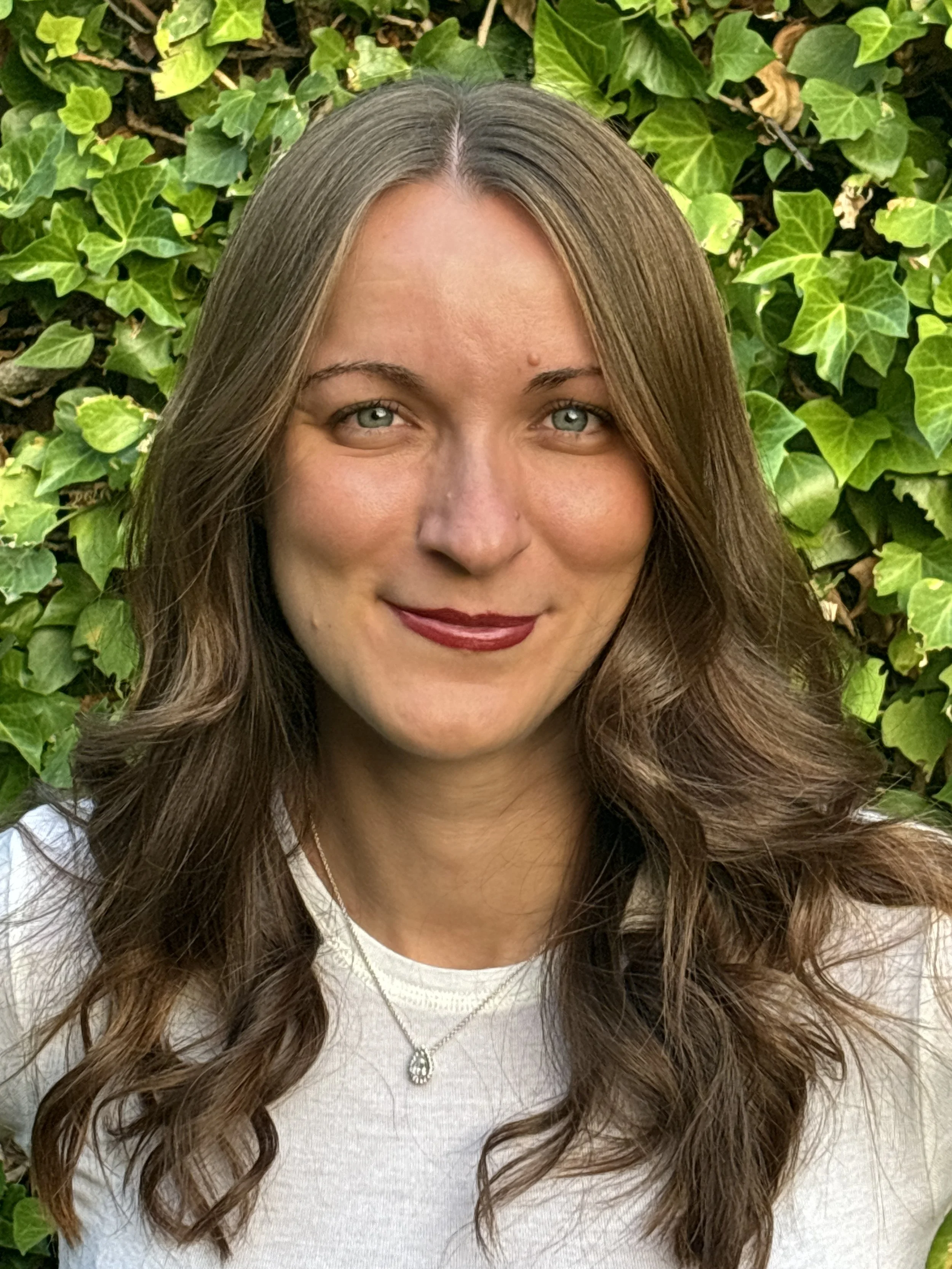 A woman with long brown hair and blue eyes smiling in front of green ivy plants.