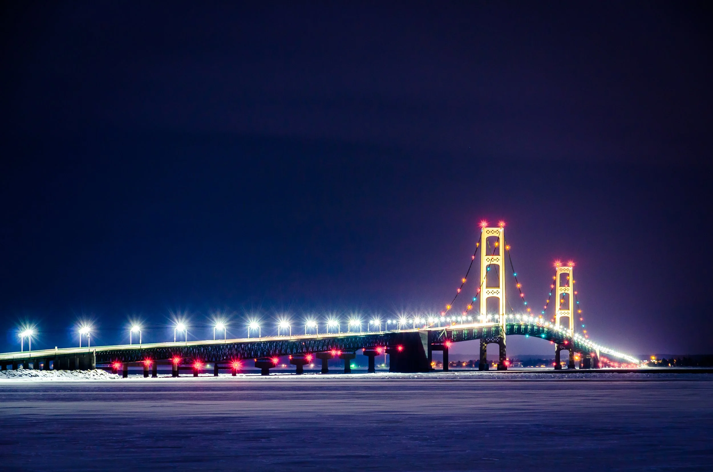 Night view of a lit-up suspension bridge with colorful lights, extending over a body of water under a dark sky.