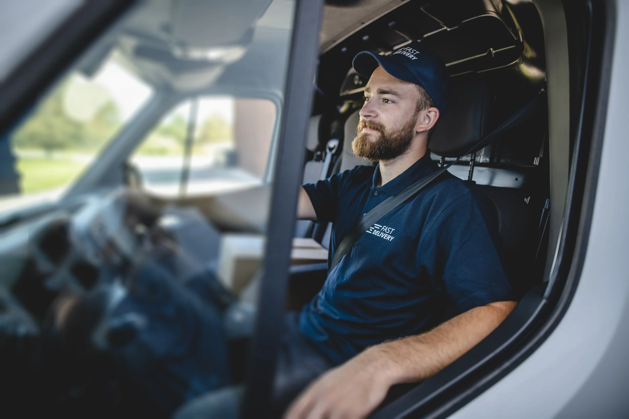 A delivery driver with a beard and wearing a navy blue cap and polo shirt sits in the driver's seat of a delivery truck, smiling and looking out the window.
