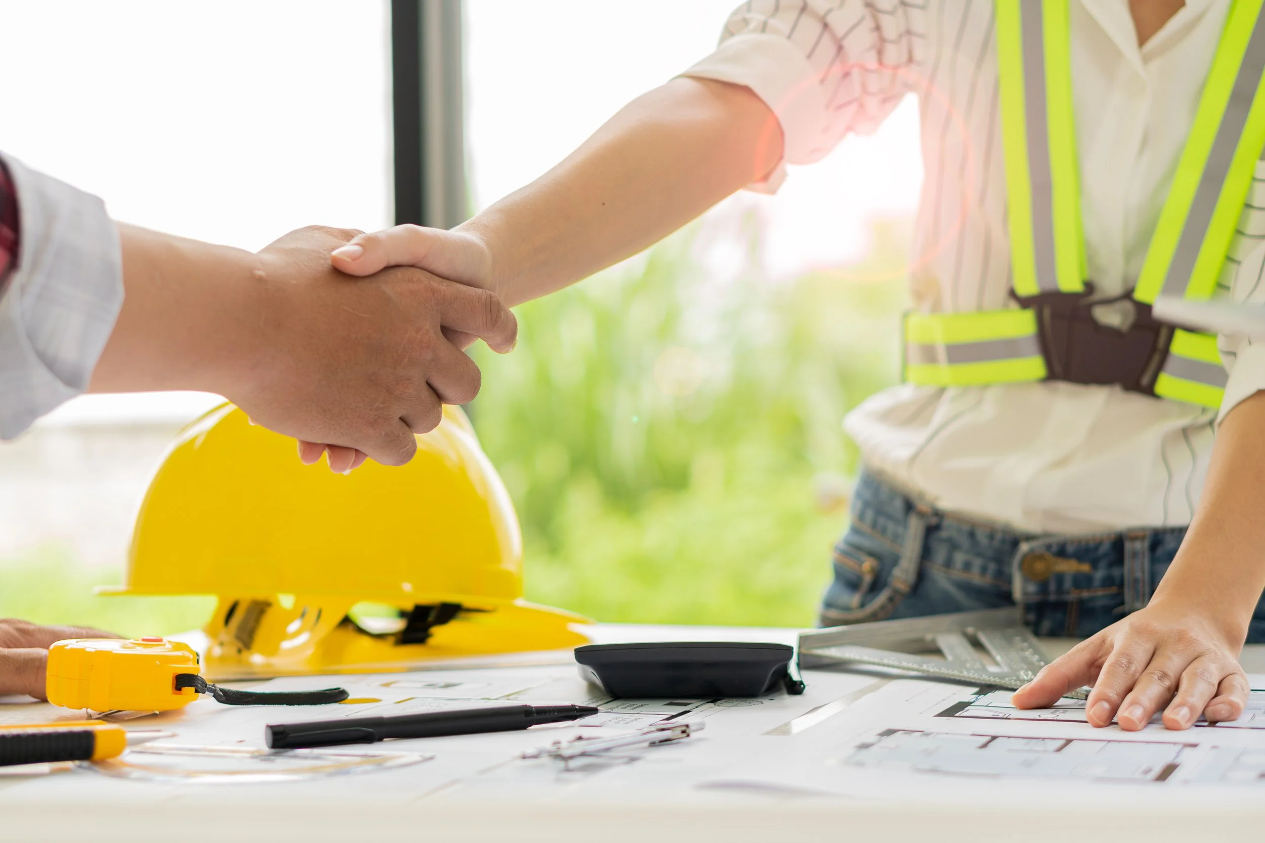 Two people shaking hands over a table with construction tools, blueprint, safety helmet, and measuring tape, suggesting a construction or business agreement setting.