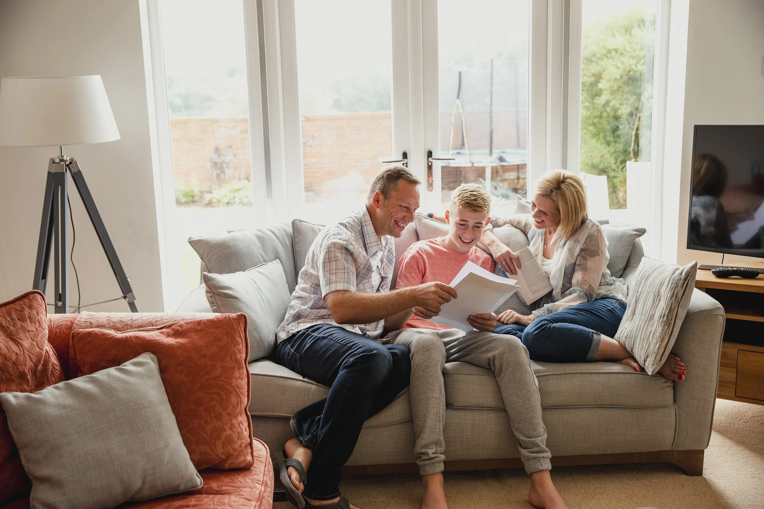 Family sitting on a sofa in a bright living room, laughing and looking at papers or photo albums together.