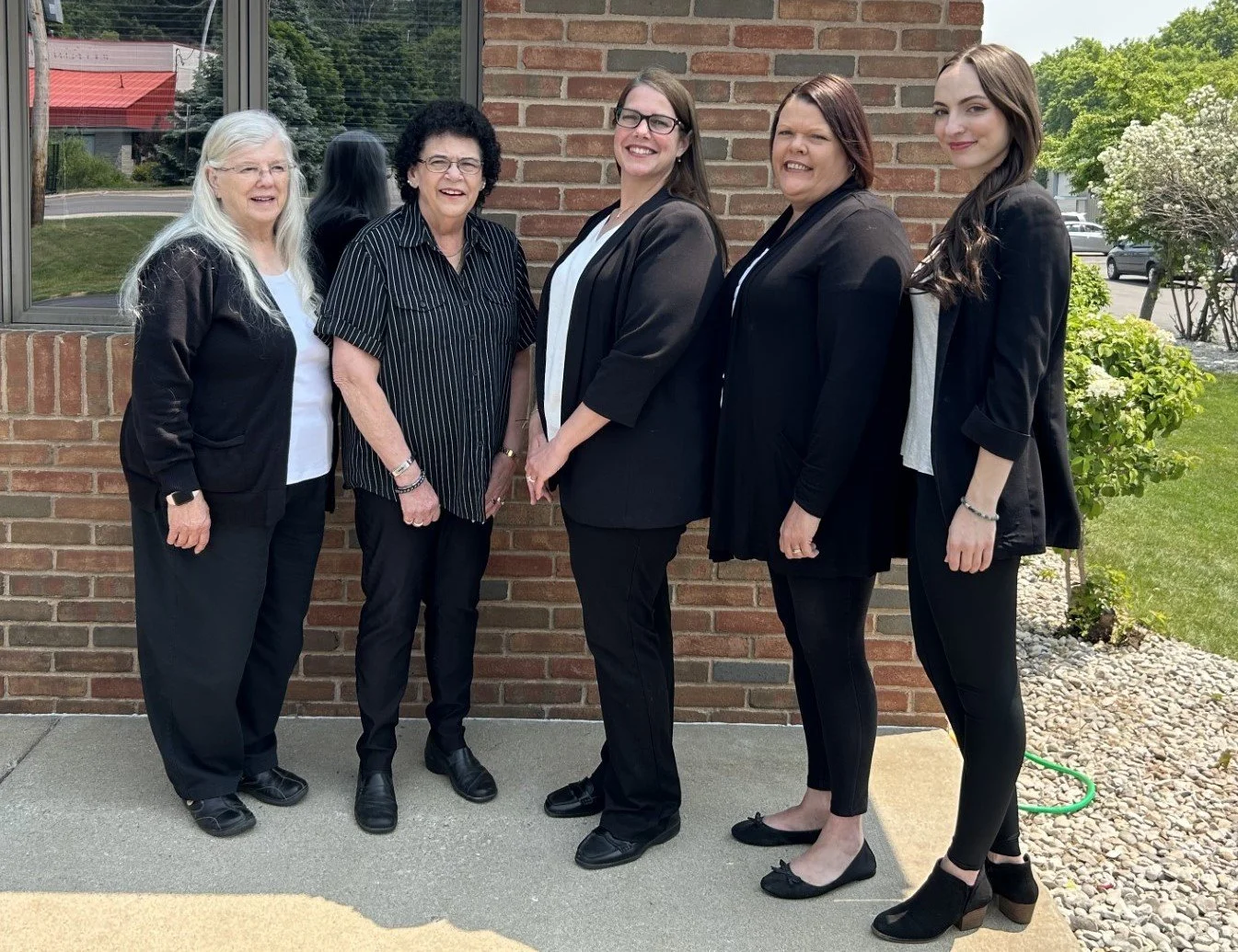Group of five women standing outside, wearing black business attire, smiling in front of a brick wall and greenery.