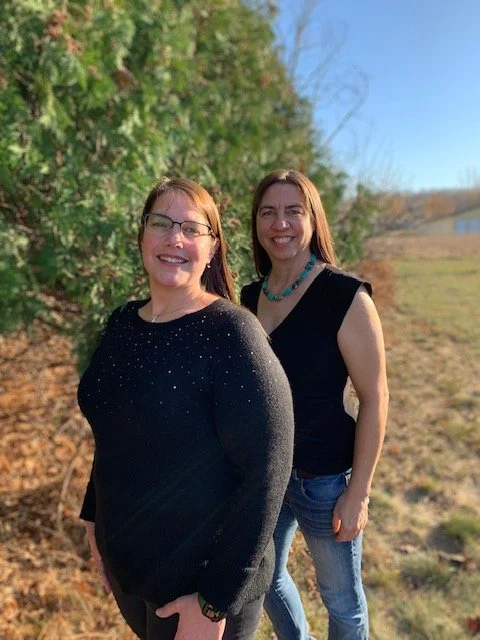 Two women standing outdoors on grass with trees and a building in the background. They are smiling at the camera, wearing casual clothing.