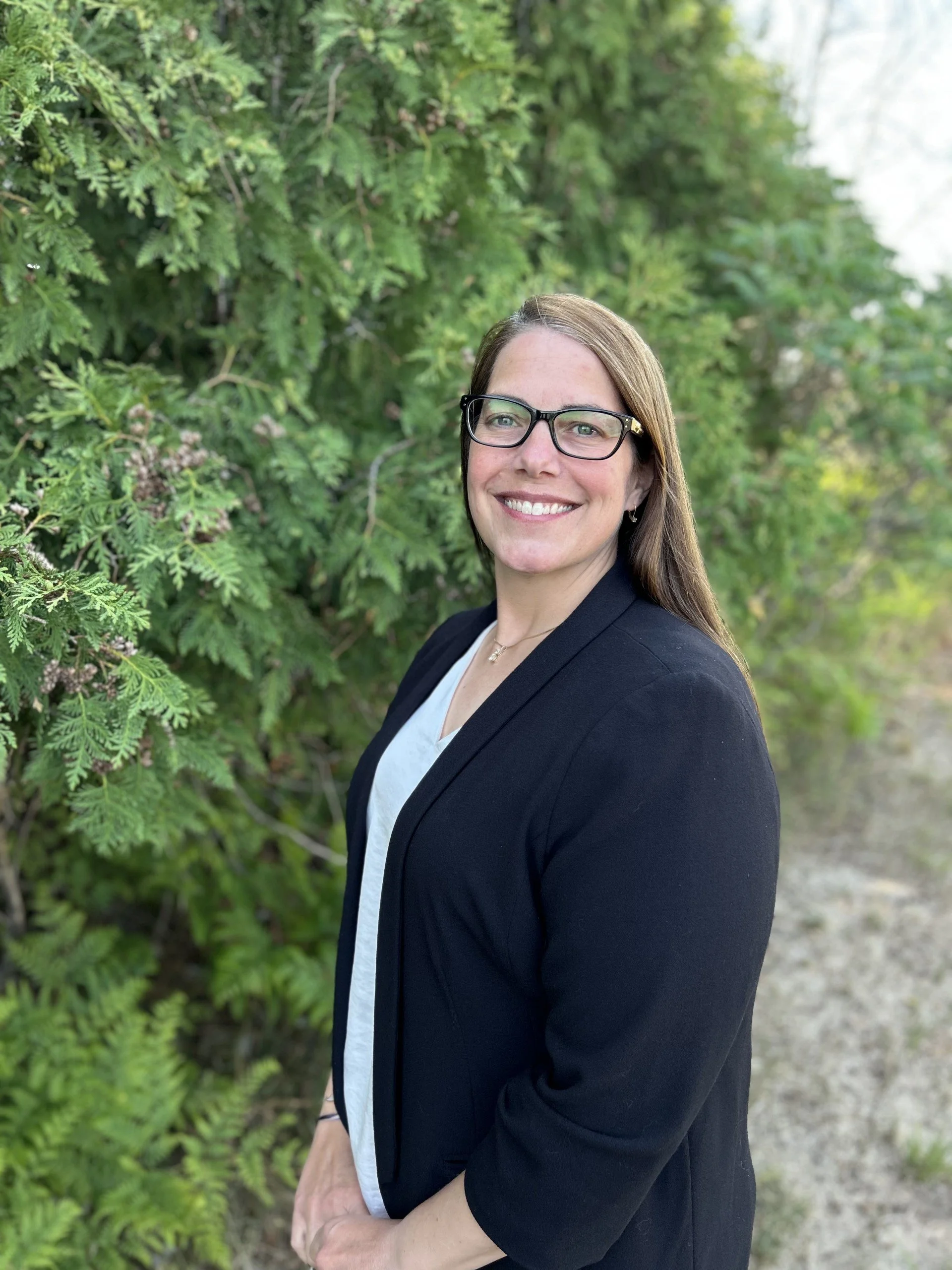 A woman with glasses and long brown hair smiling outdoors near green foliage, wearing a black blazer and white top.
