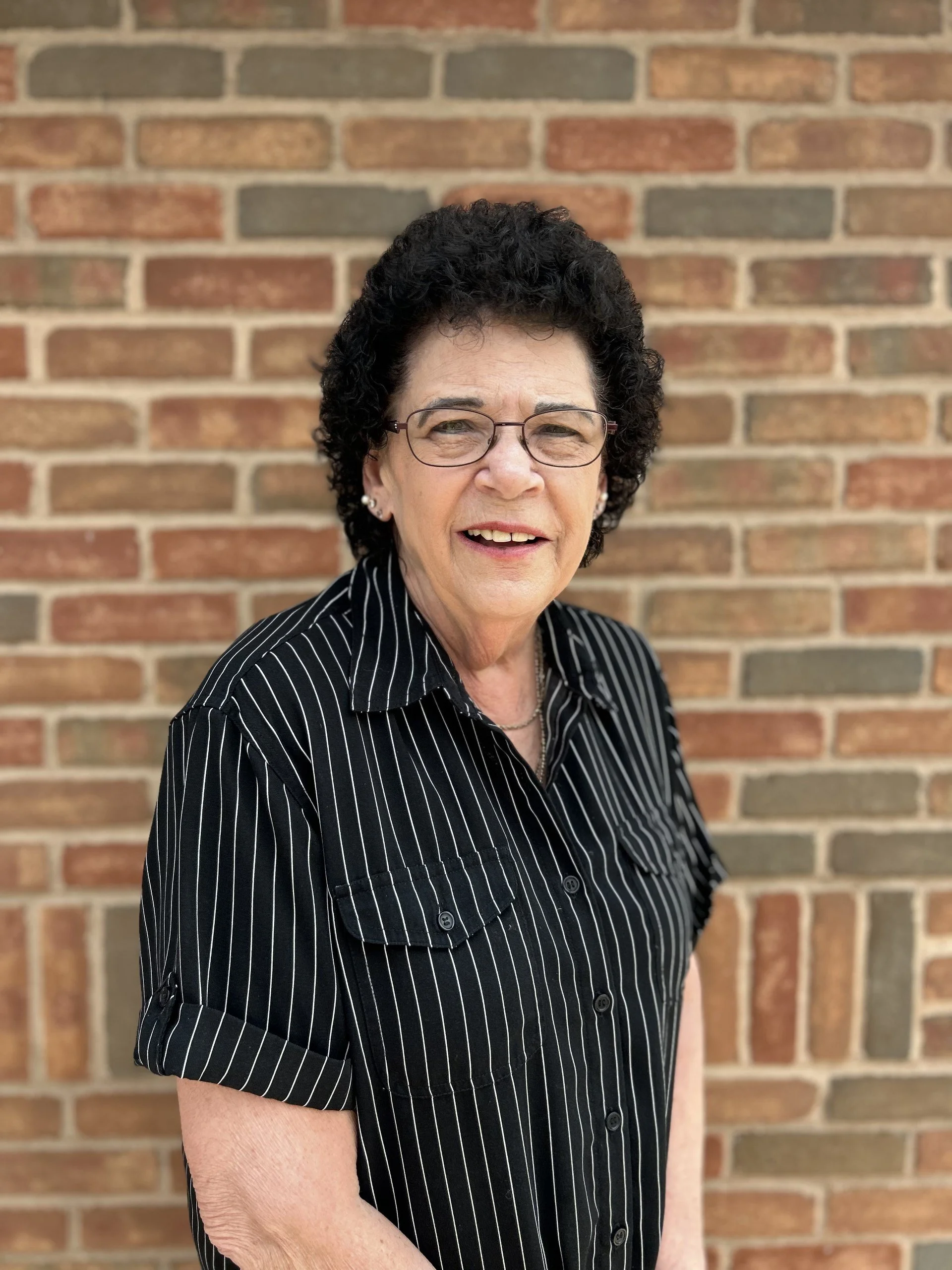 A woman with curly dark hair and glasses standing in front of a brick wall, wearing a black and white pinstripe shirt.