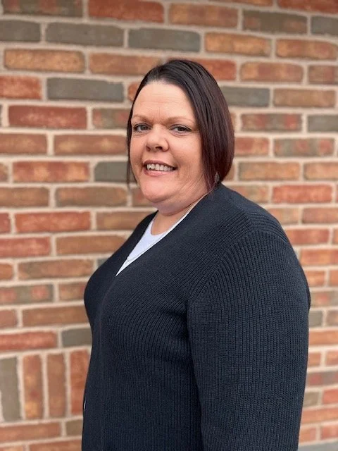 A woman with dark brown hair and light skin, smiling, standing against a brick wall, wearing a dark sweater over a white shirt.