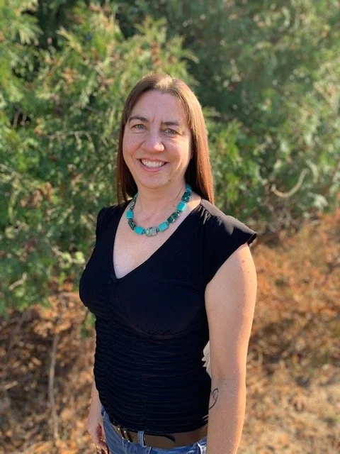 A woman smiling outdoors with green foliage in the background, wearing a black top and turquoise necklace.