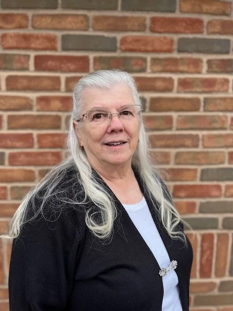 An elderly woman with long gray hair wearing glasses, a black jacket, and a white shirt standing outdoors in front of a brick wall.